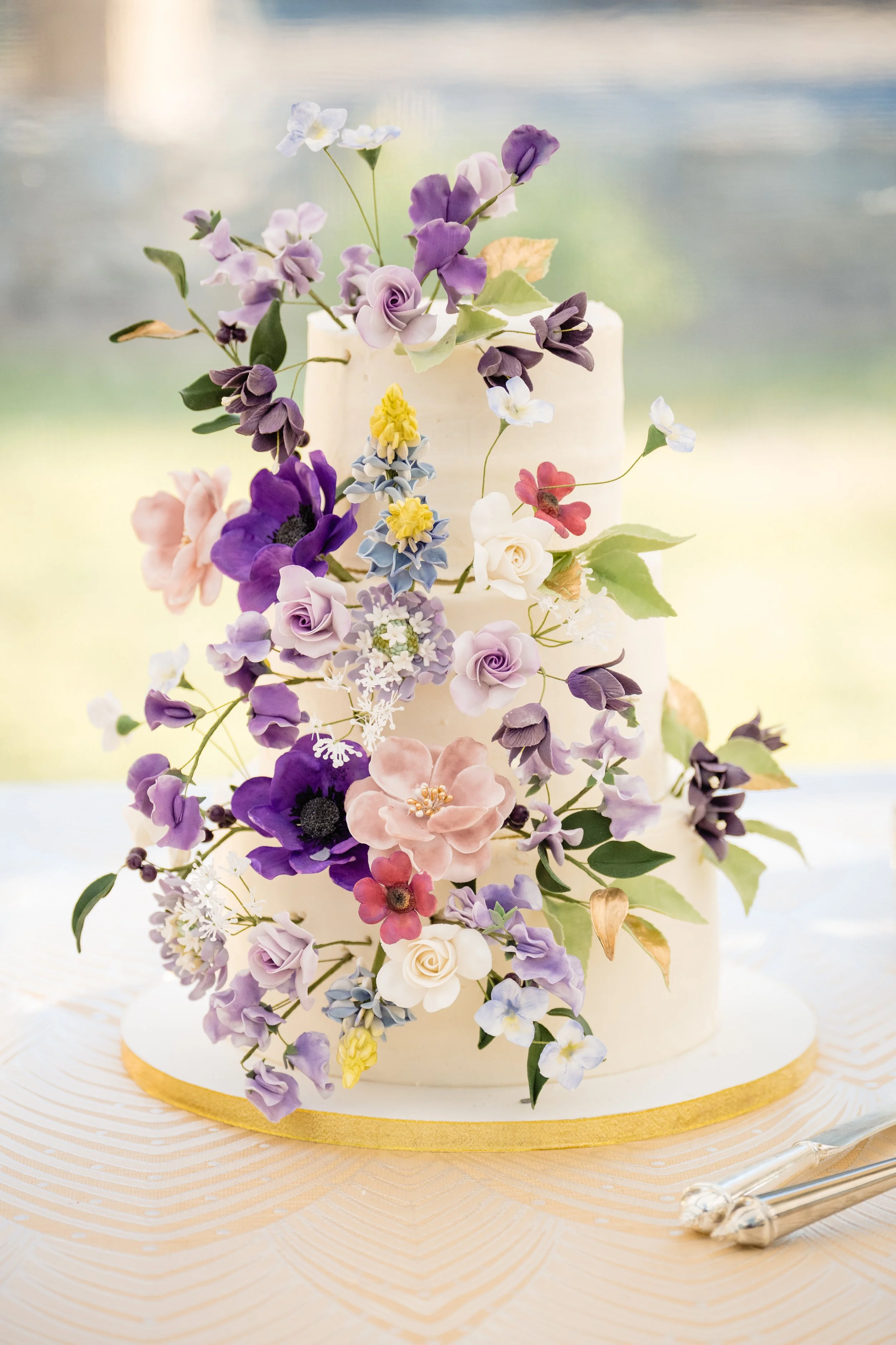 A multi-layer wedding cake decorated with an array of pastel-colored and purple flowers and green leaves, with a gold trim around the base, placed on a white patterned tablecloth with silver cake serving utensils beside it.