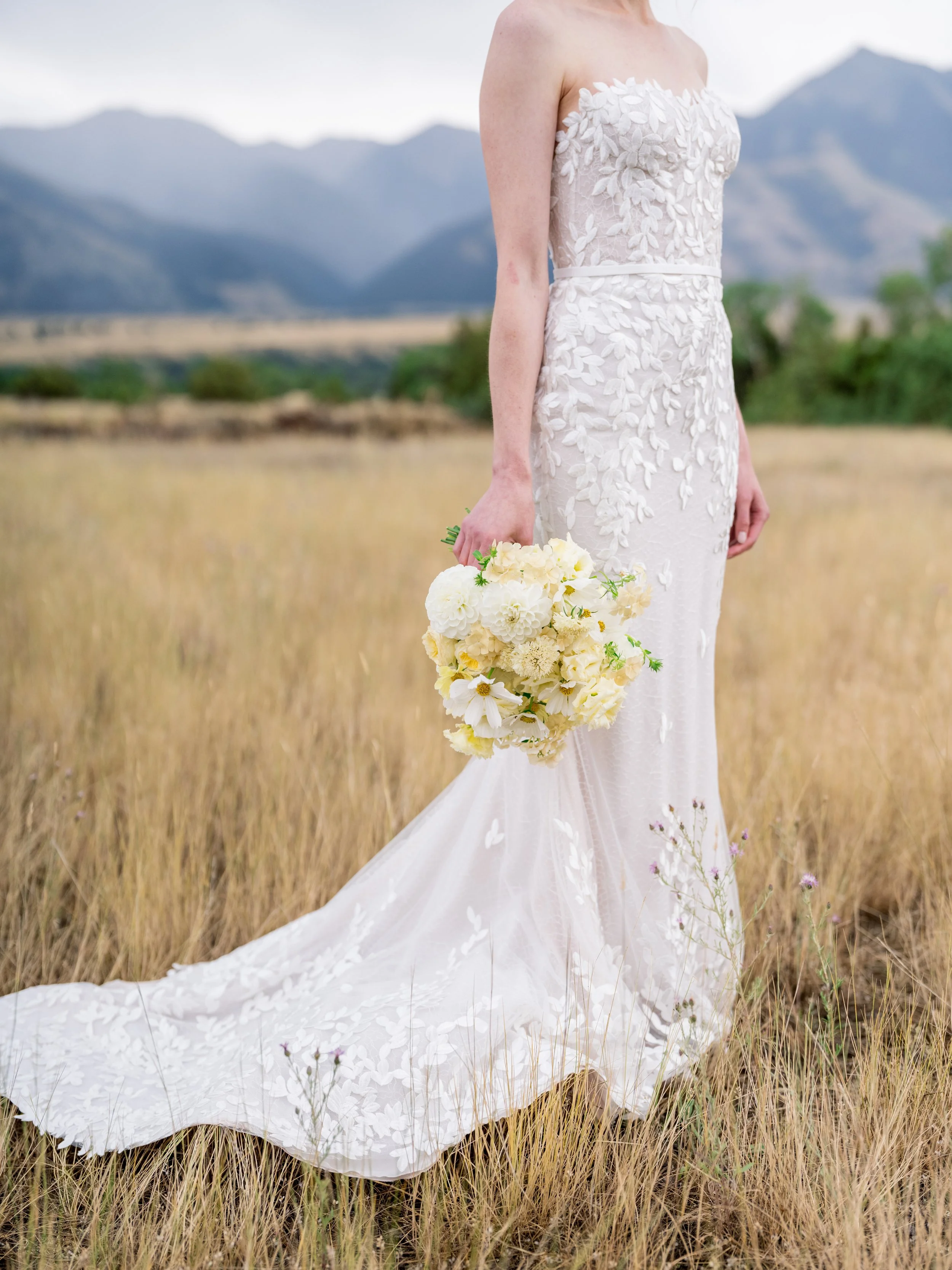 Bride in a fitted wedding gown with floral embroidery holding a white bouquet in a Montana field with mountain views