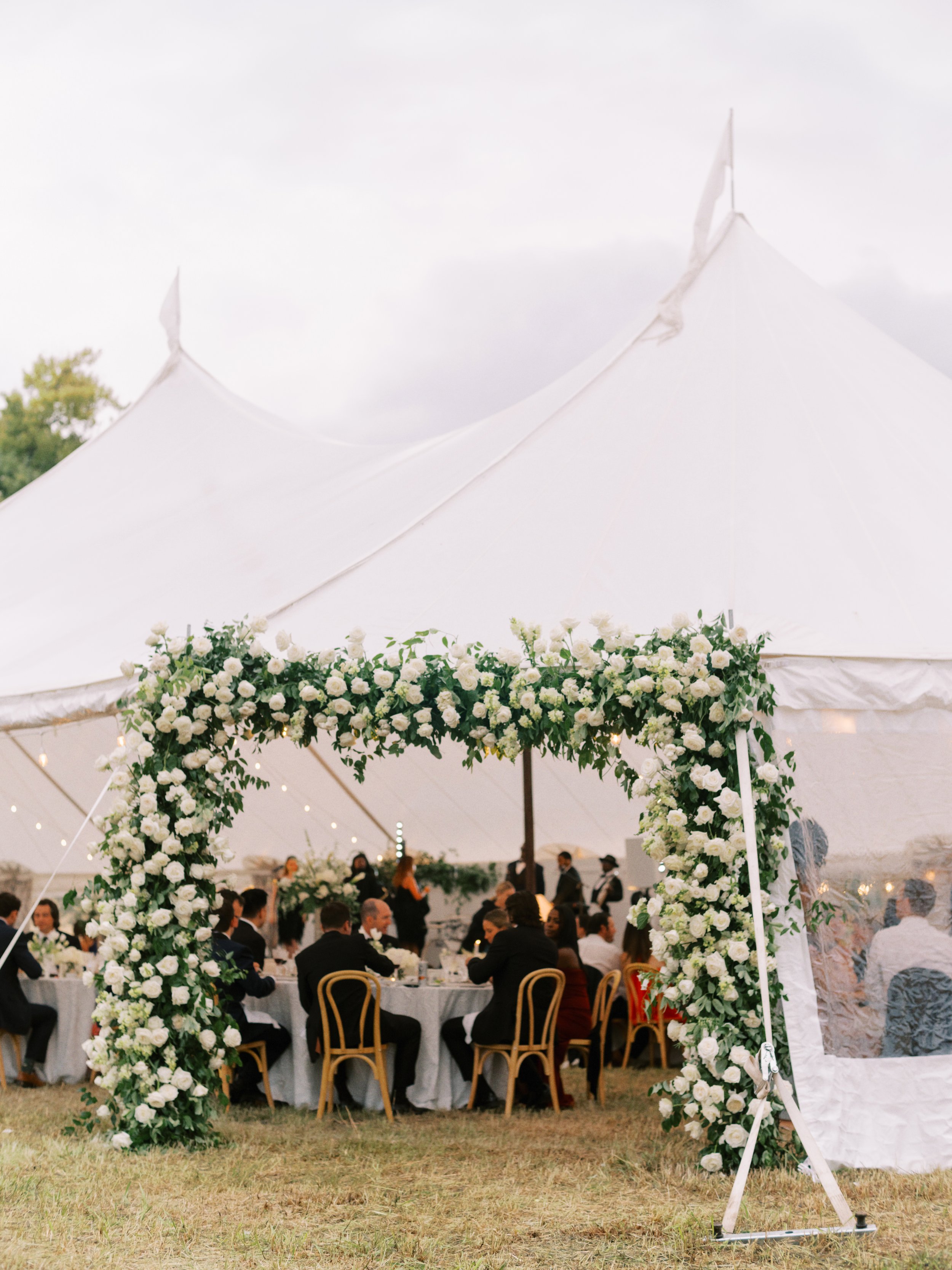 A wedding reception under a large white tent decorated with an arch of white flowers and greenery, with guests sitting at tables and mingling inside.