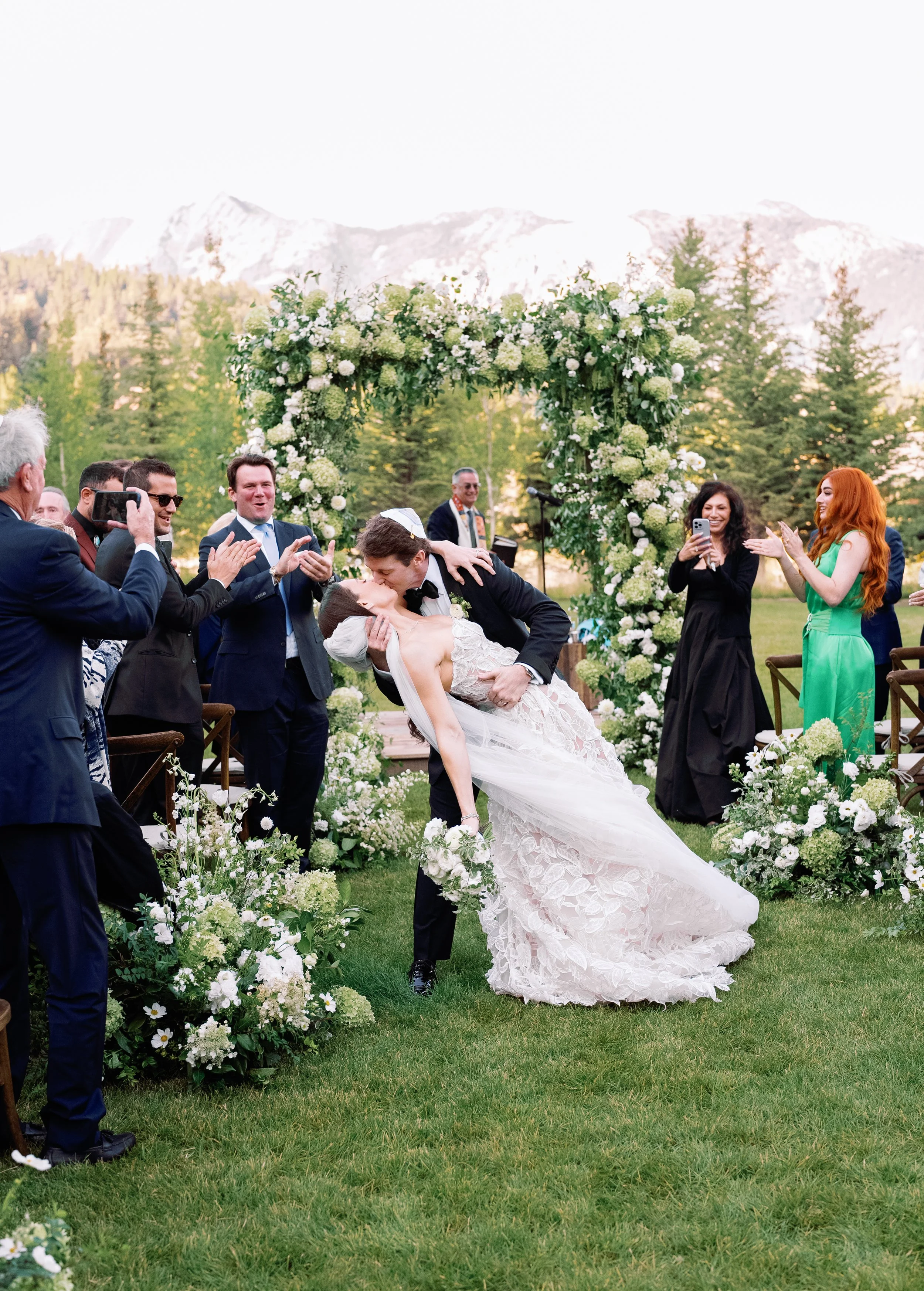 A wedding ceremony outdoors with a bride and groom kissing, surrounded by guests and a floral arch, with mountains in the background.