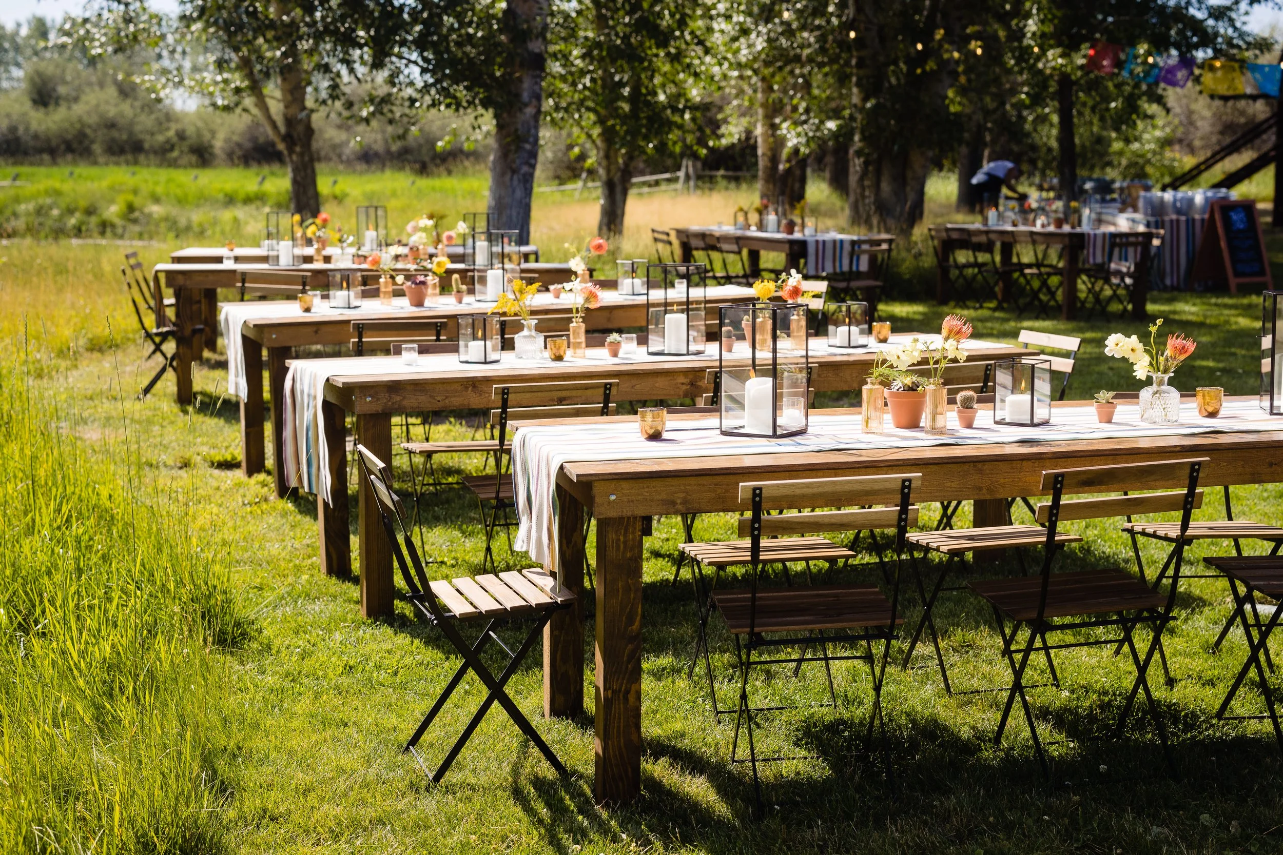 Outdoor wedding or event setup with long wooden tables decorated with flowers, candles, and lanterns, under trees in a green field on a sunny day.