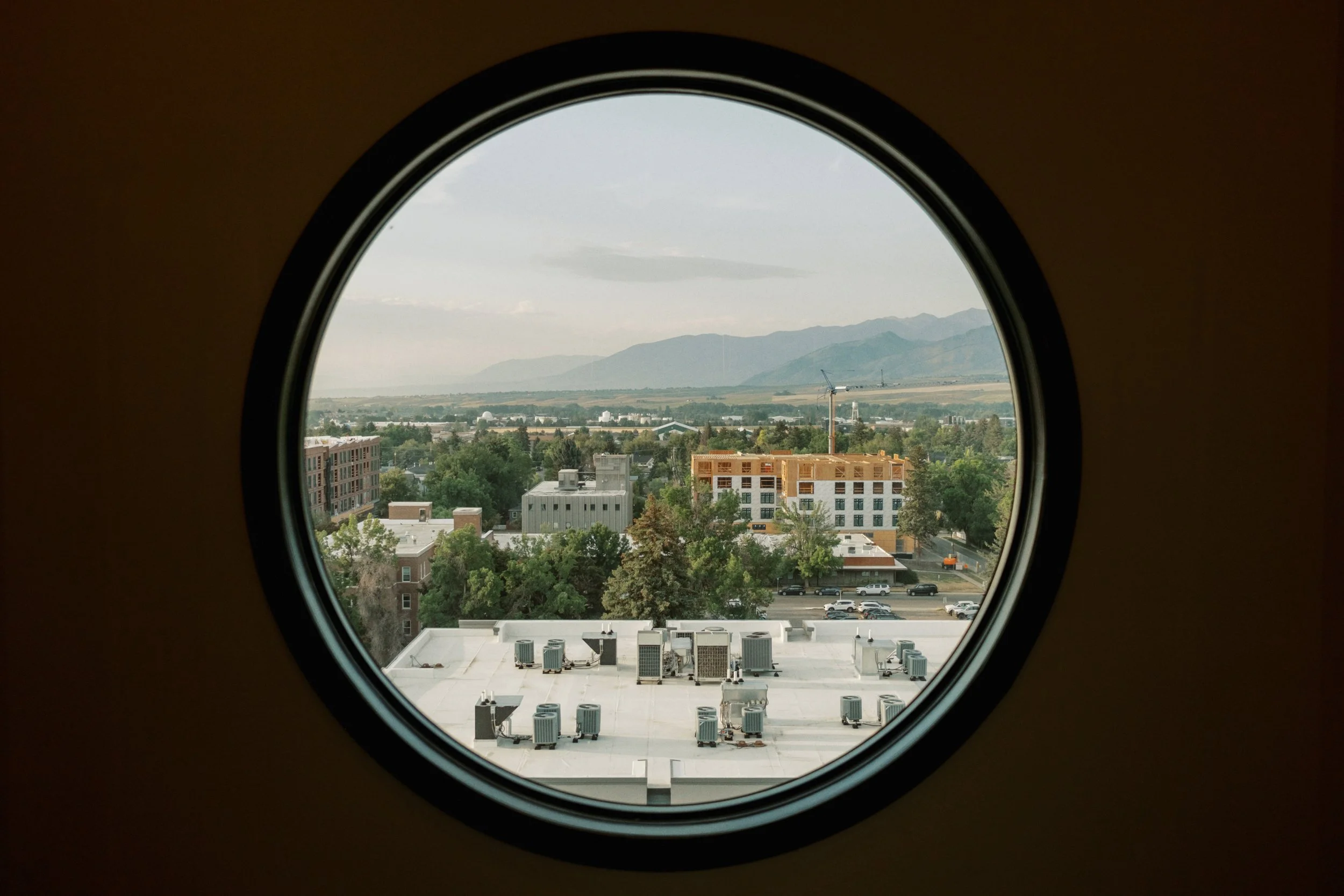 View of a cityscape with trees, buildings, and mountains in the distance, seen through an oval airplane window from a rooftop.
