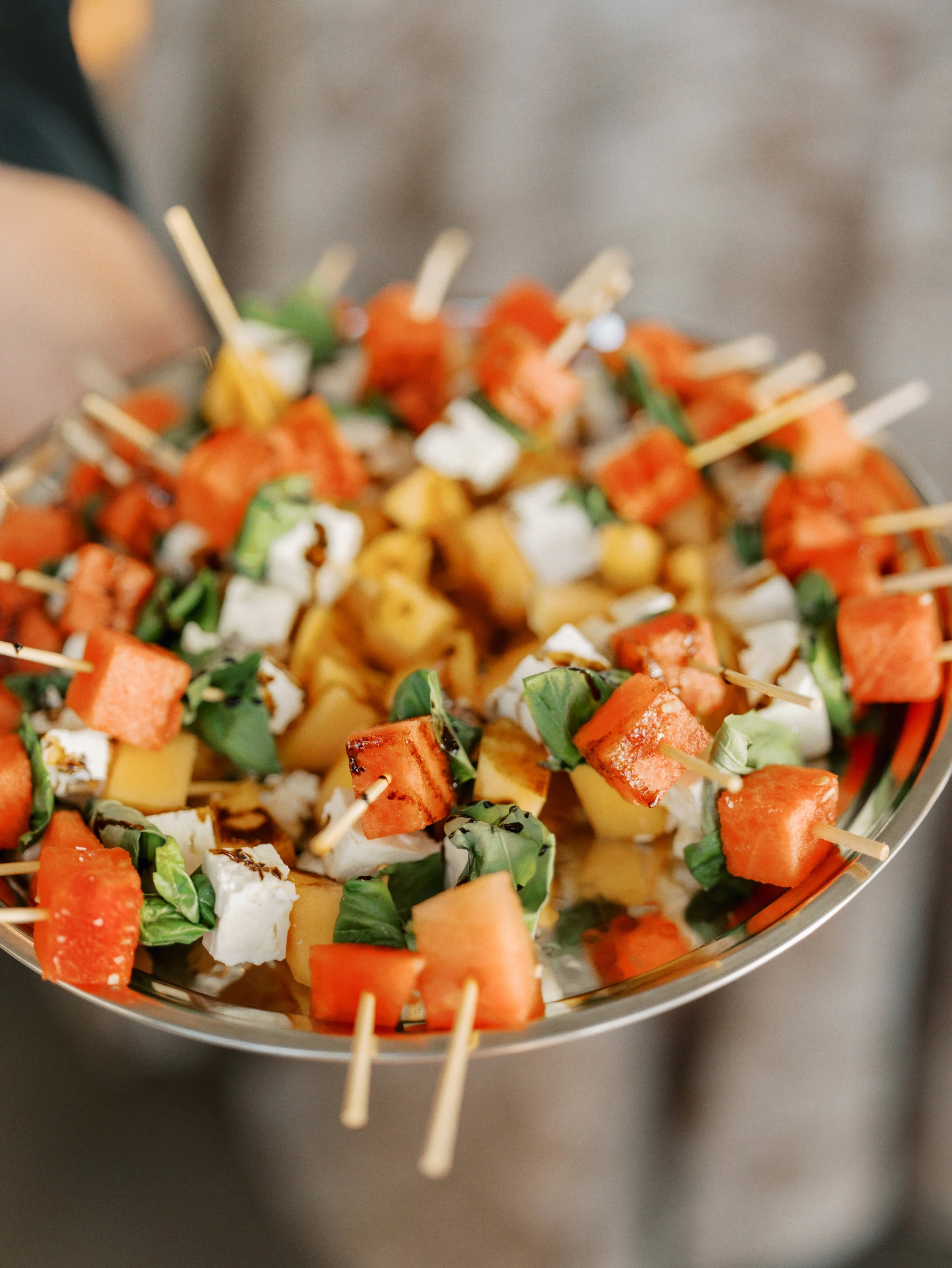 Close-up of a tray of assorted skewered appetizers with pieces of watermelon, cheese, basil, and grilled vegetables.