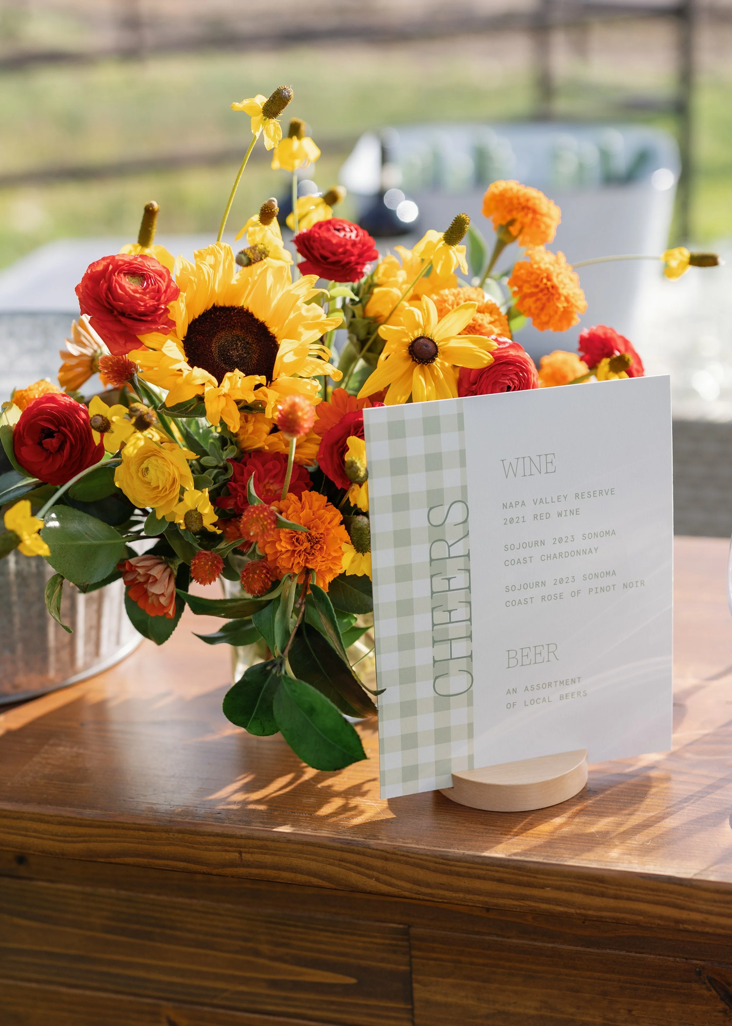 Vibrant sunflower and dahlia floral arrangement in warm orange and yellow tones beside a marble wedding sign, by Wild Blume