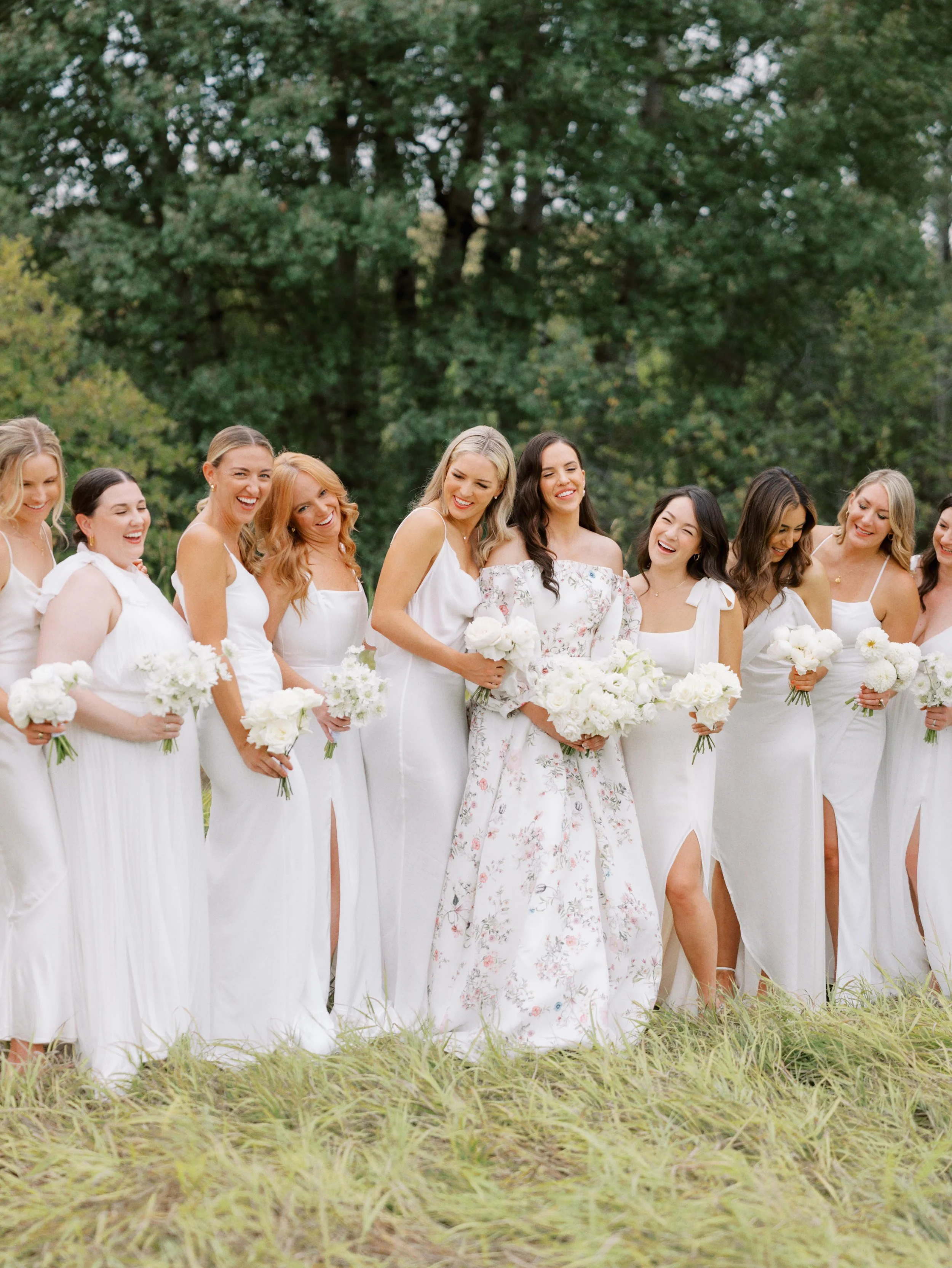 Group of women in white dresses holding white flower bouquets, standing outdoors in a grassy area with trees in the background.