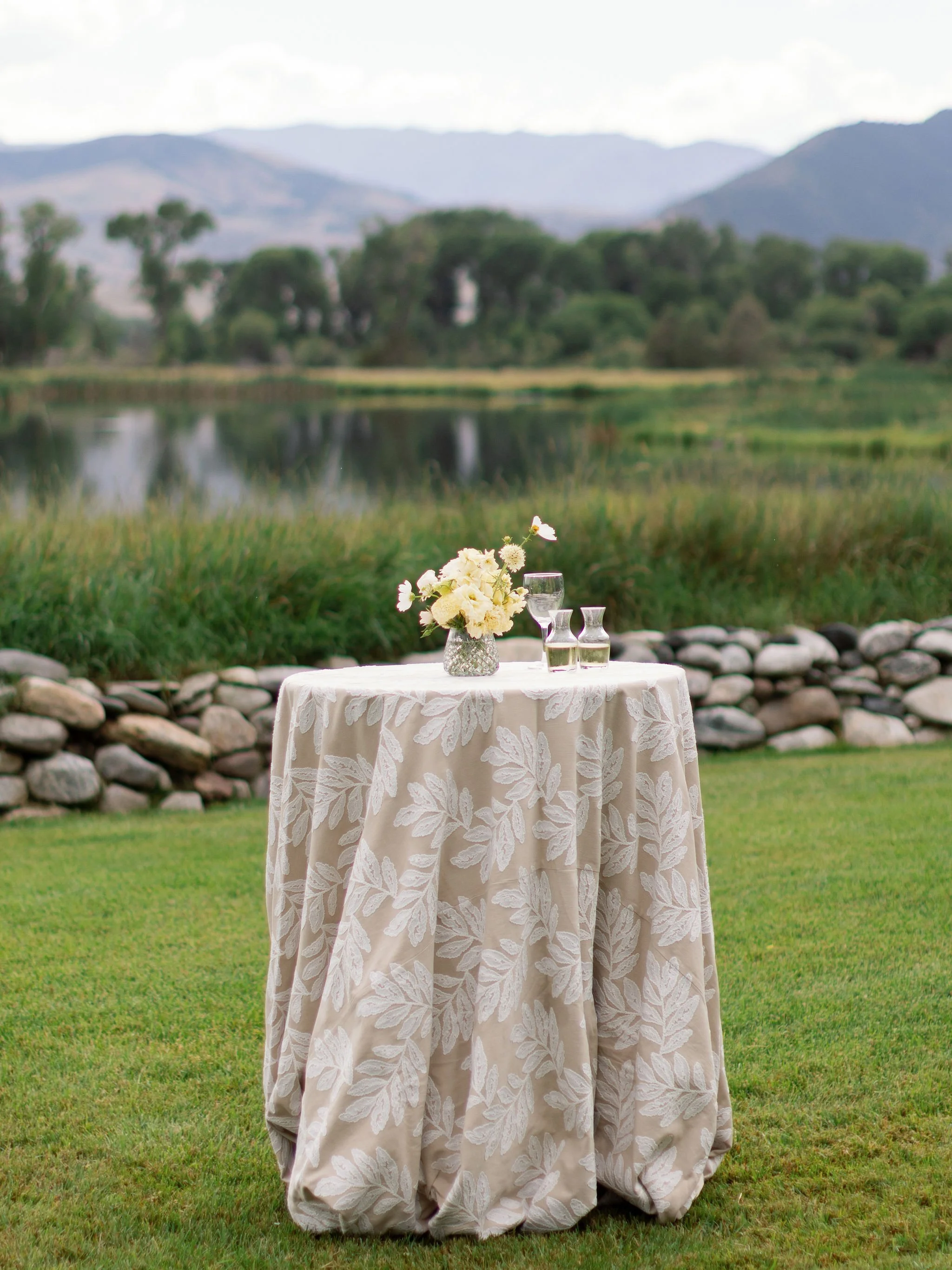A round table with a cream-colored tablecloth decorated with a floral pattern, set outdoors on grass with a scenic background of mountains, trees, and a river, and decorated with a glass vase of white and light yellow flowers and three glass bottles.