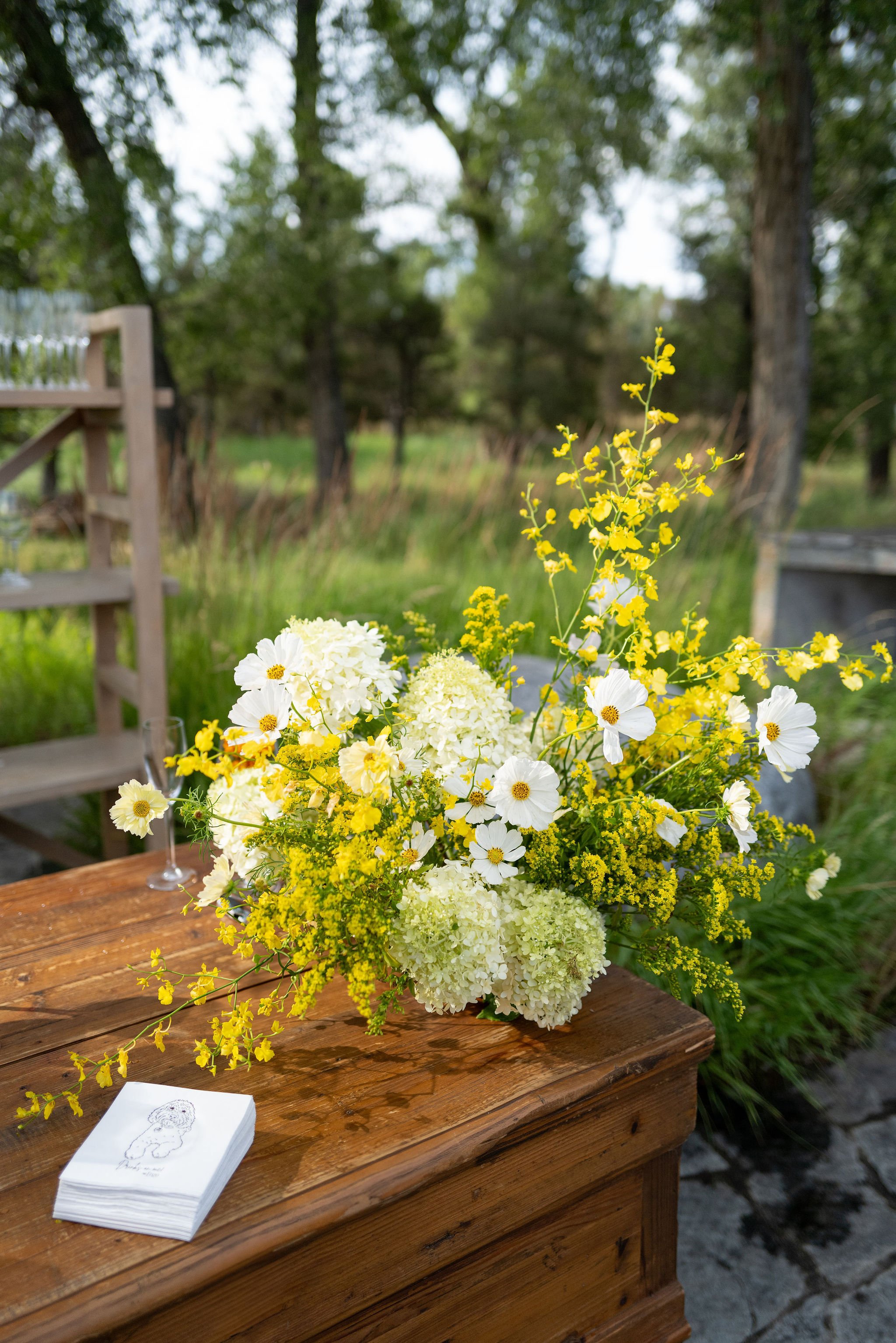 A bouquet of white and yellow flowers on a wooden table outdoors, with a blurred background of trees and greenery.
