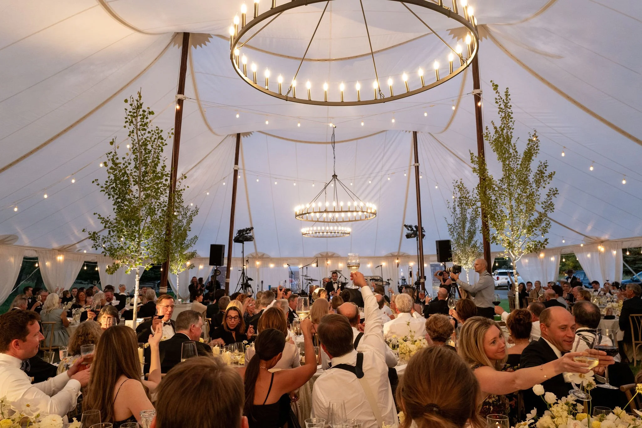 A large indoor event under a white tent with elegant chandeliers and string lights. Many guests in formal attire are seated at tables, enjoying the dinner, while a band performs on stage in the background.