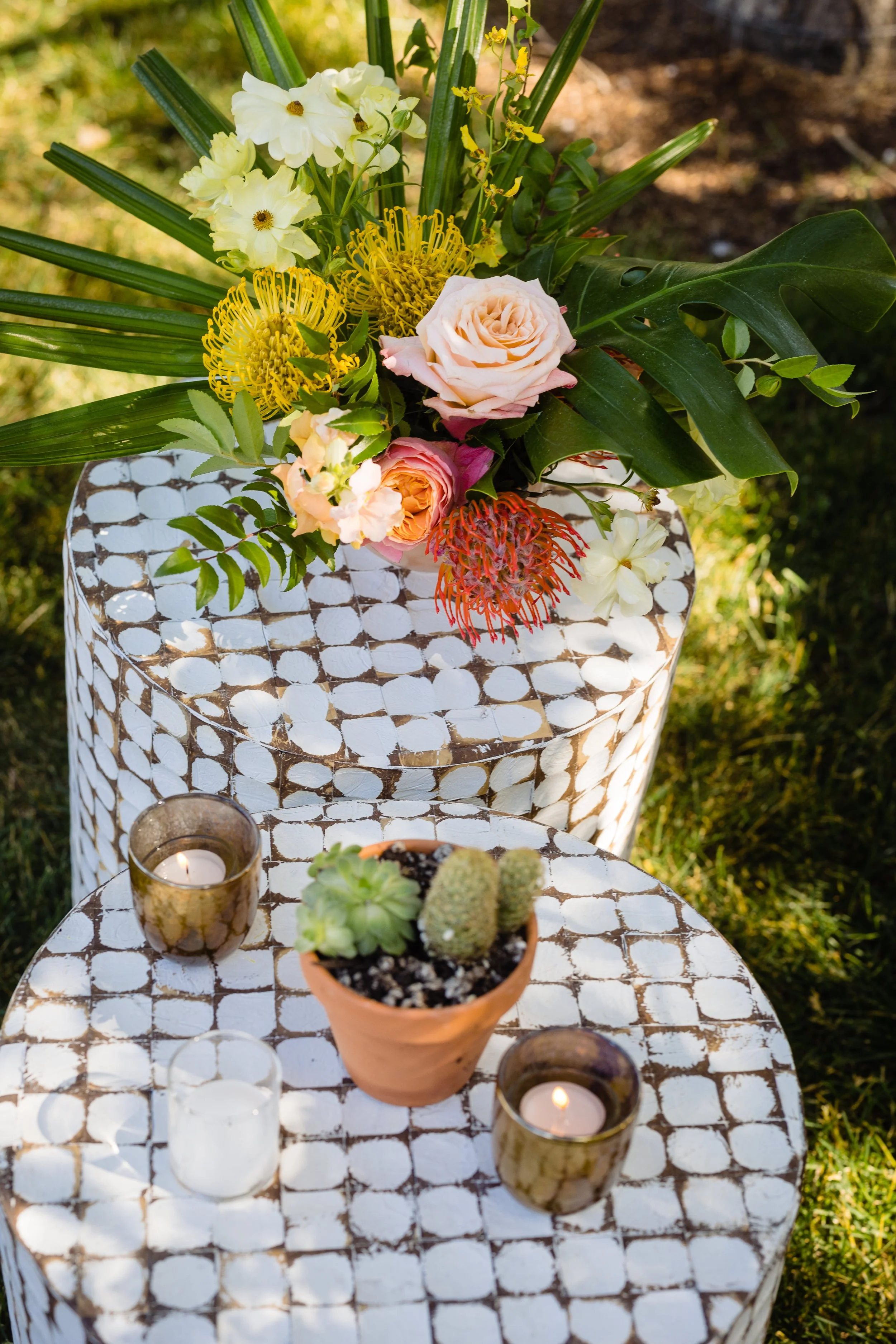 A floral arrangement with roses, lilies, and tropical leaves on a white patterned table. Candles and a small cactus in a terracotta pot are on the table, set outdoors on grass.