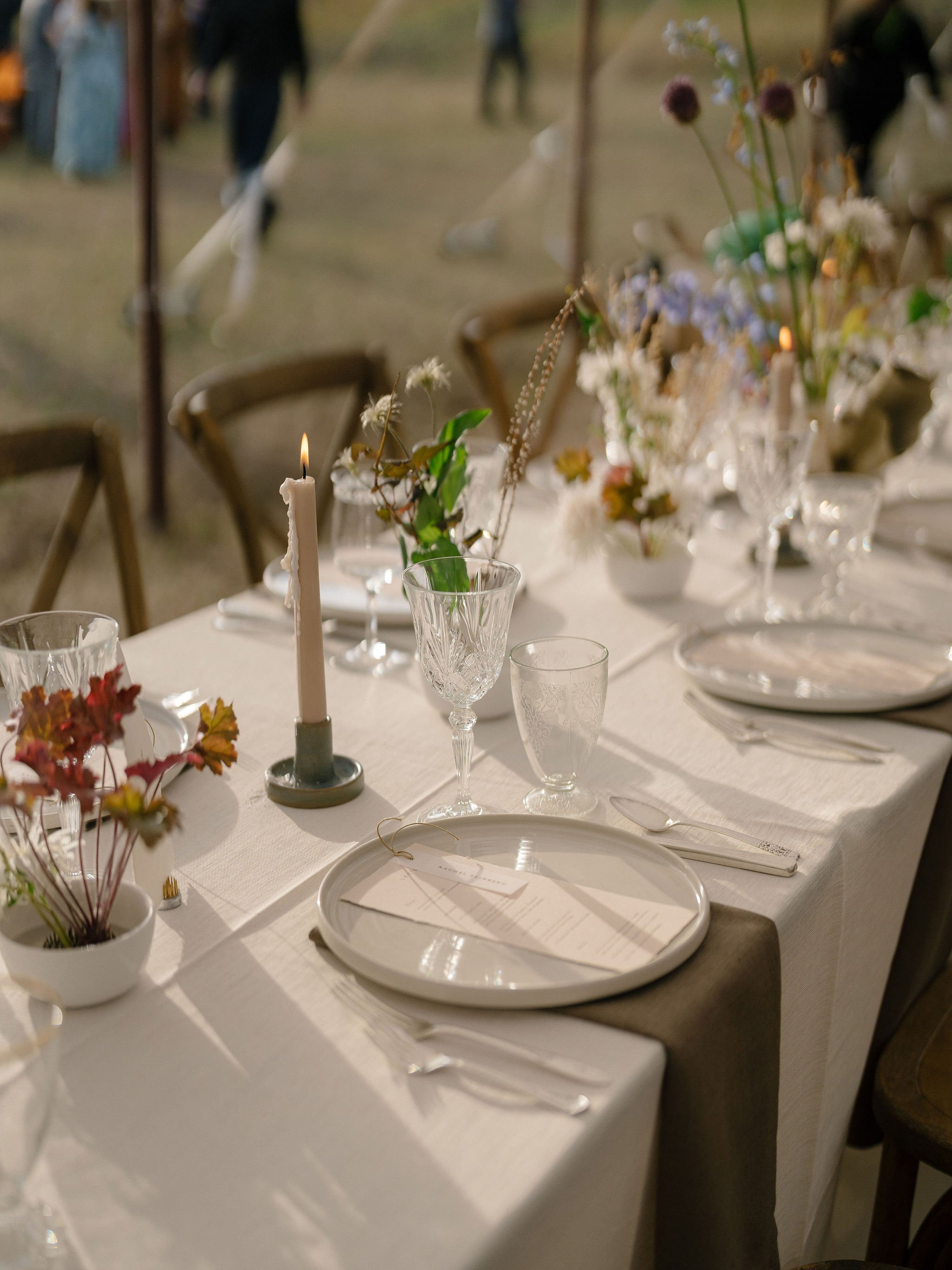 A decorated banquet table with flower arrangements, glassware, a lit candle, and place settings, set in a softly lit environment, possibly at a wedding or event.