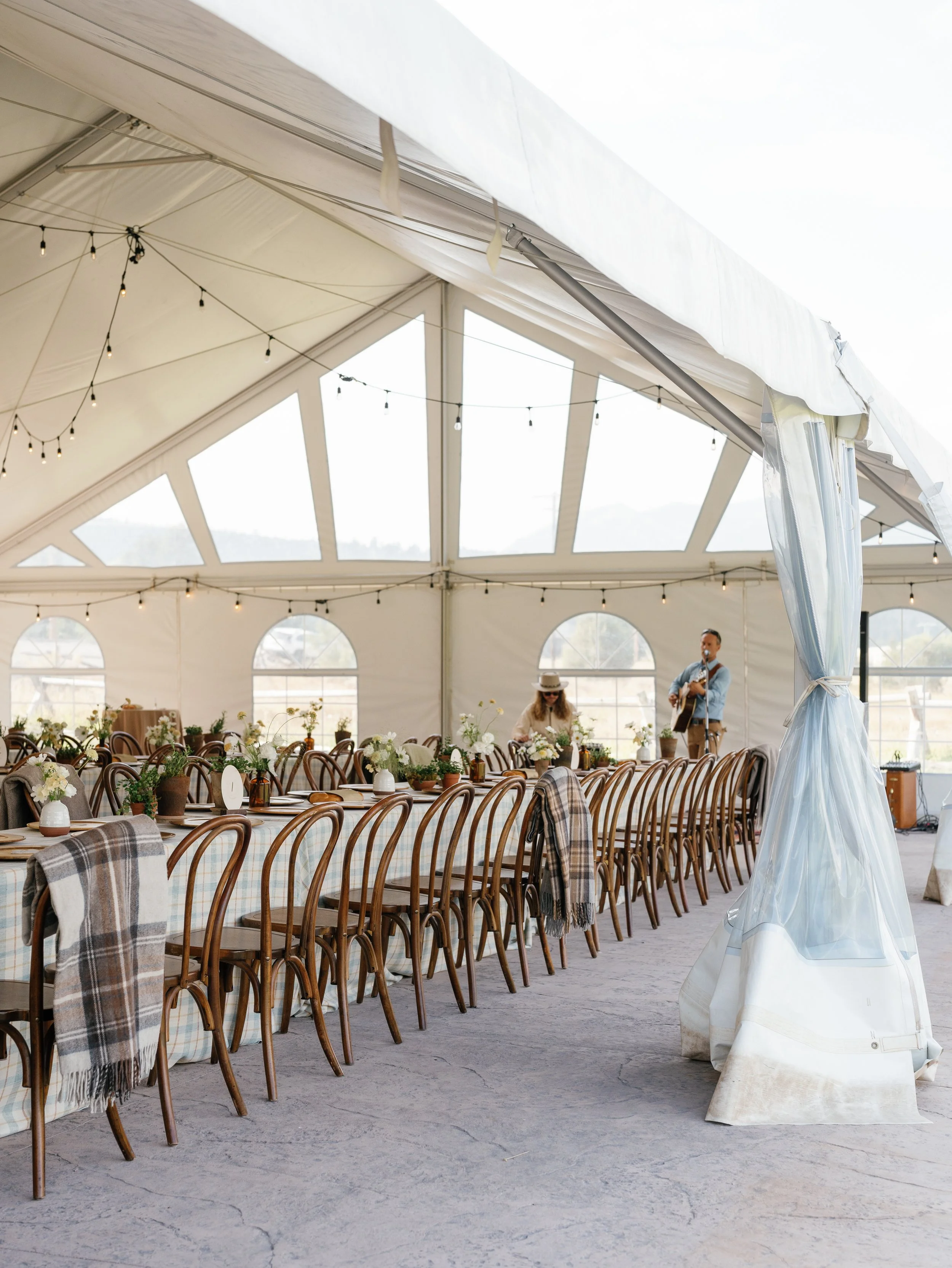 Inside a large white event tent with string lights hanging from the ceiling. Surrounding a long table with wooden chairs, some with plaid shawls draped over them. Decor includes small plant pots and flowers, with some people visible in the background