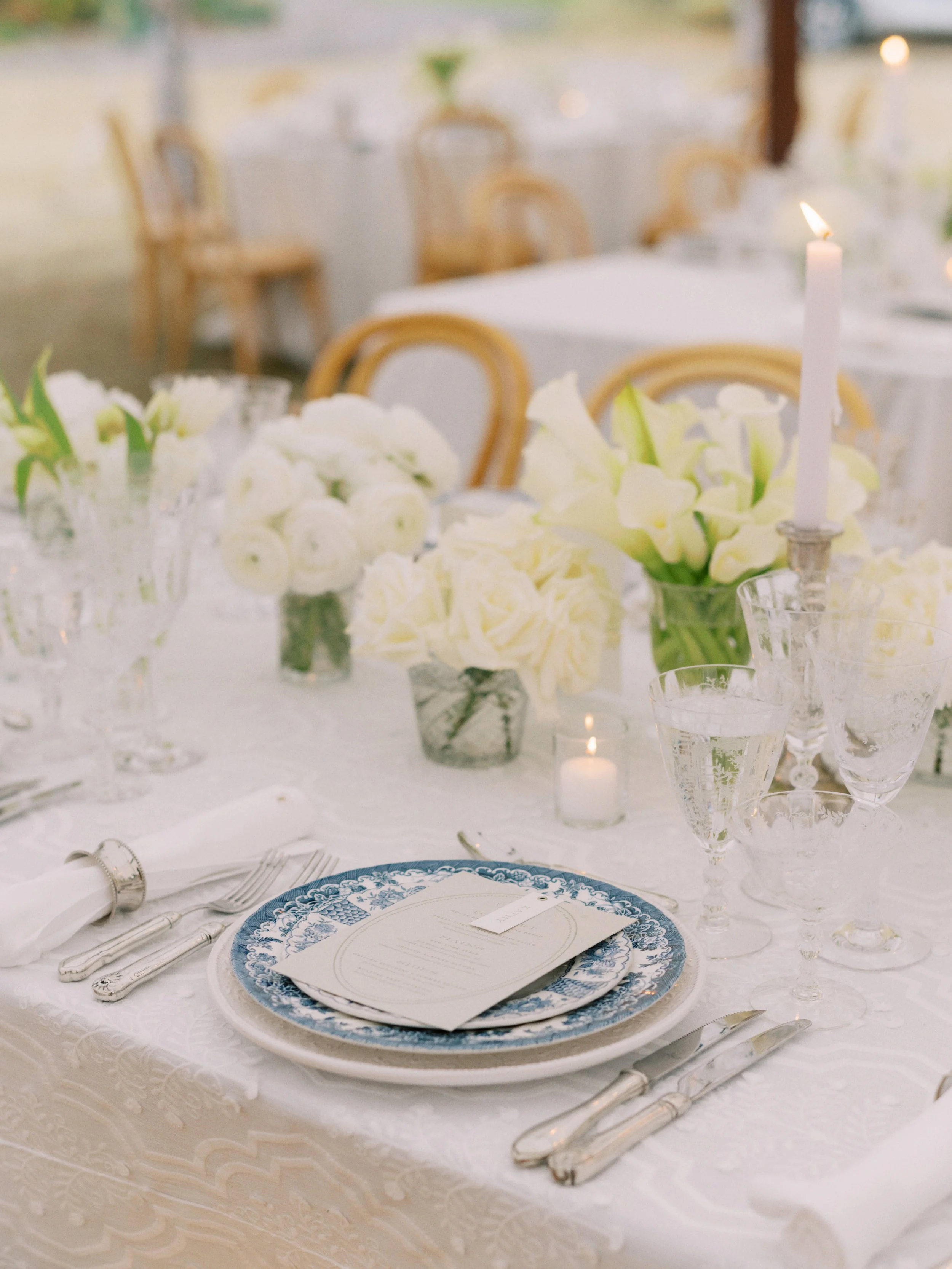 Elegant table setting with blue and white patterned plate, menu, silverware, glassware, floral centerpiece, and white candle on a white tablecloth, likely for a wedding or formal event.