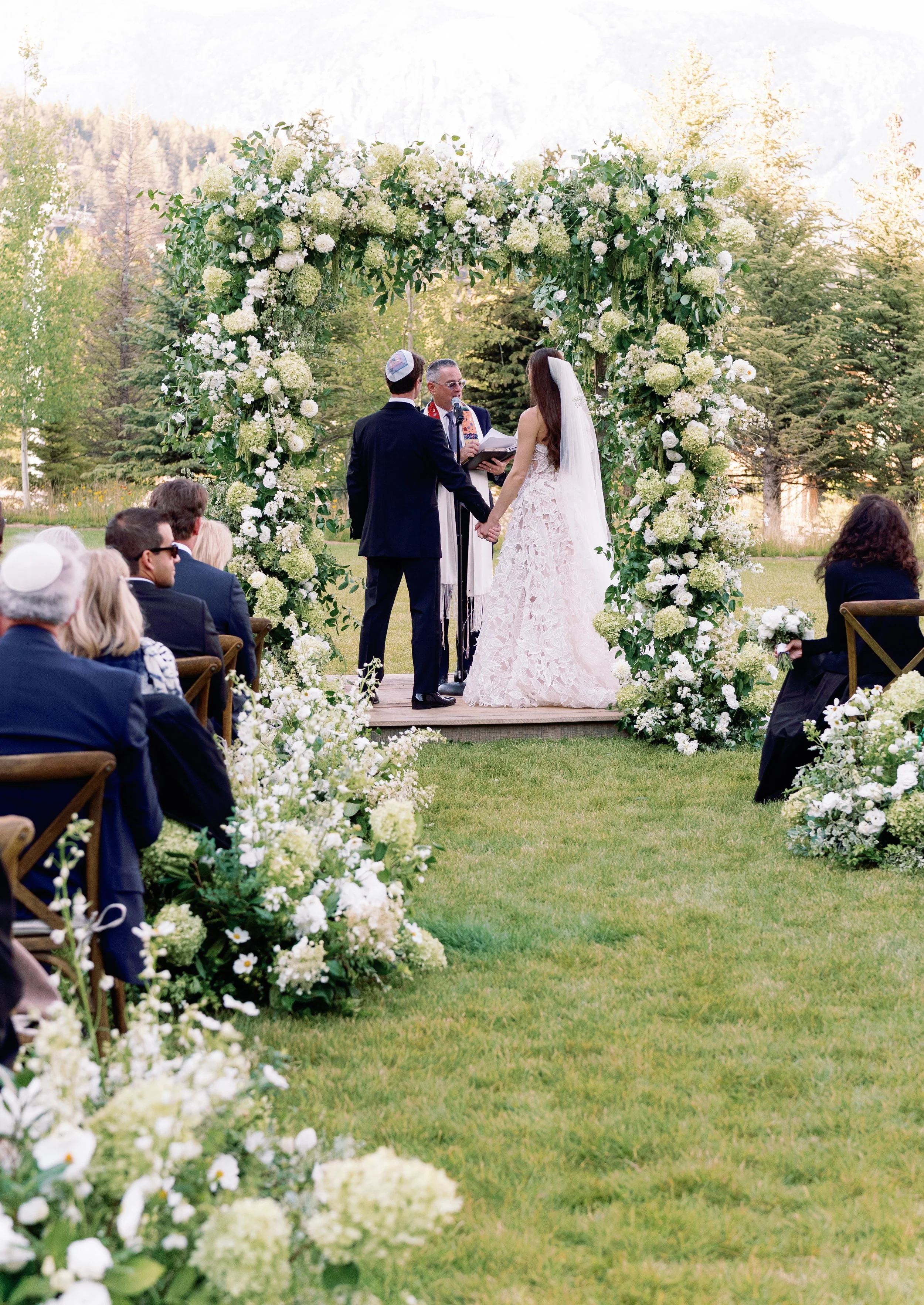 A wedding ceremony taking place outdoors with a bride and groom holding hands beneath a floral arch. The officiant is speaking into a microphone. Guests are seated on either side, dressed in formal attire, with trees and greenery in the background.