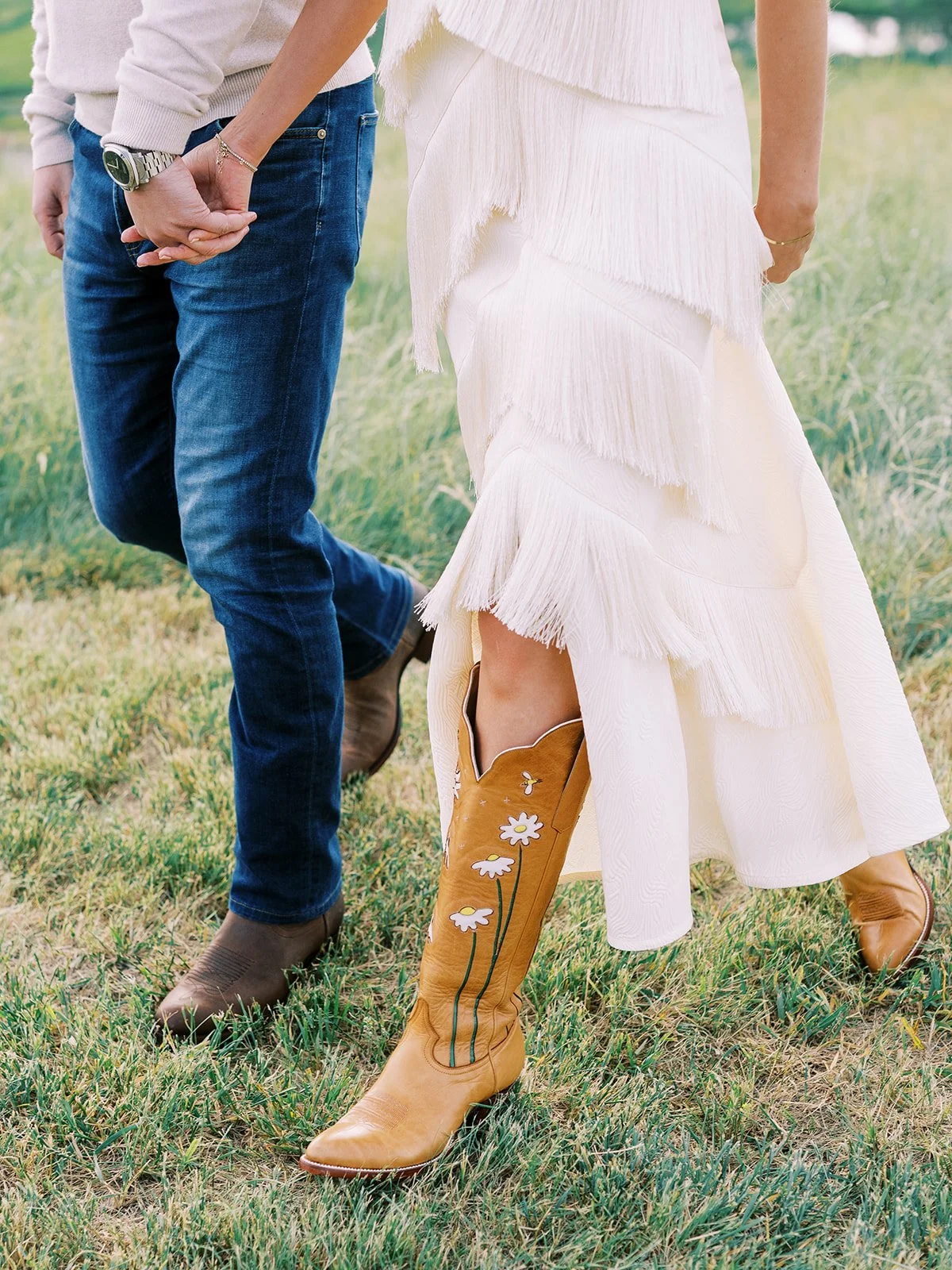 A couple holding hands while standing on grass, with the woman wearing cowboy boots with floral embroidery, and the man dressed in jeans and boots.