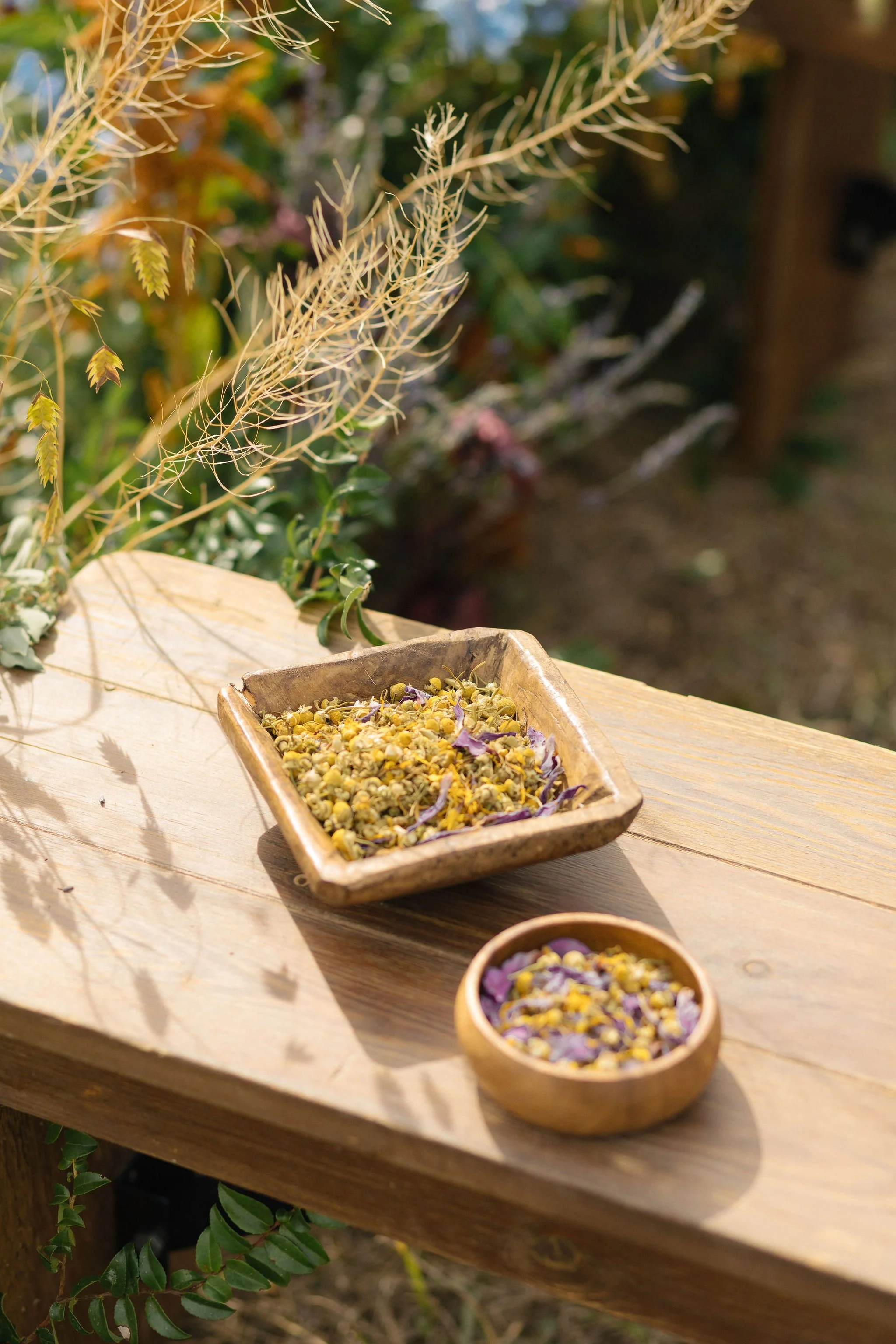 A wooden table outdoors with dried flowers and herbs in small wooden and ceramic bowls, surrounded by greenery.