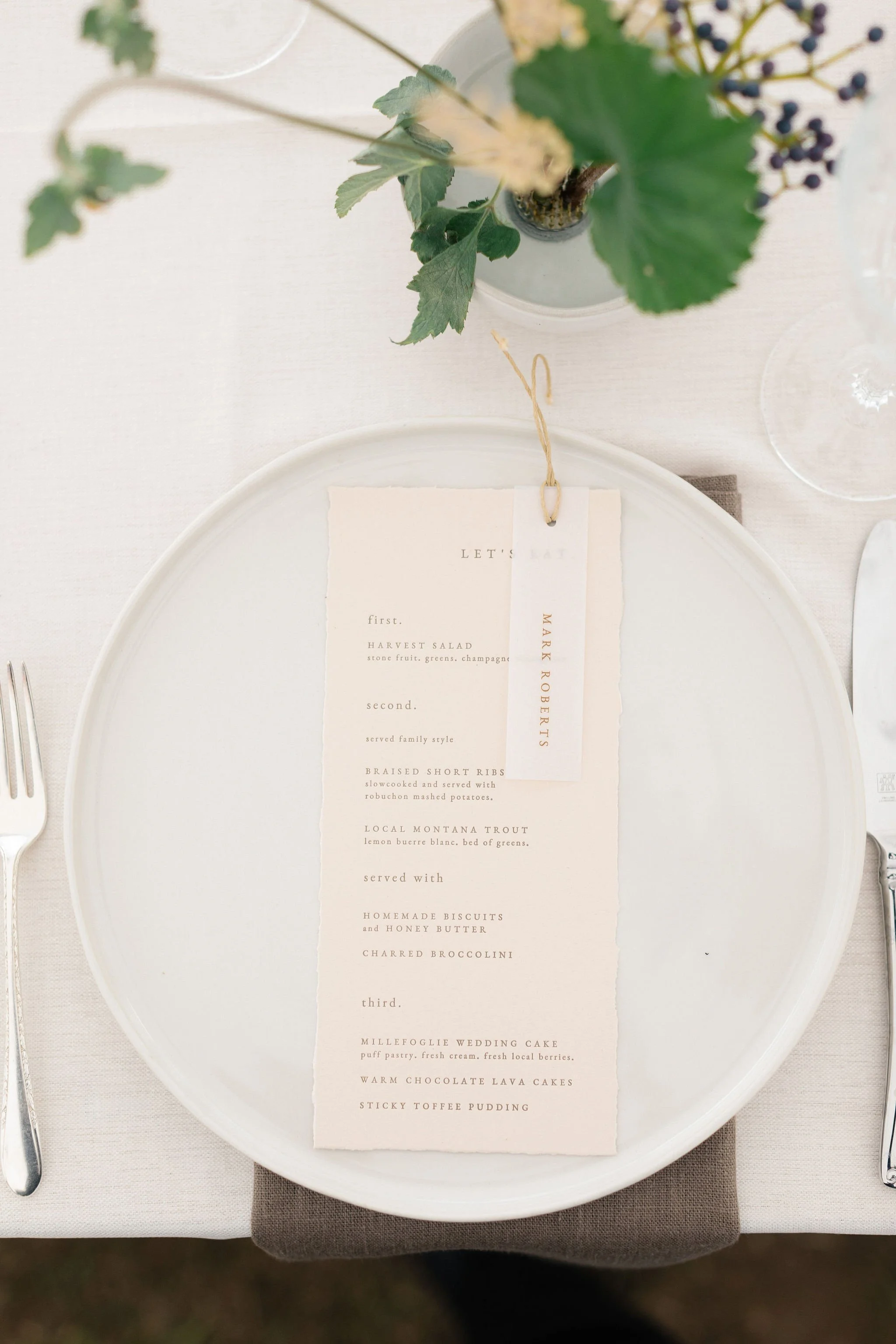 A wedding menu on a white plate with a floral centerpiece and glass of water, set on a table with a white tablecloth and a brown napkin underneath the plate.