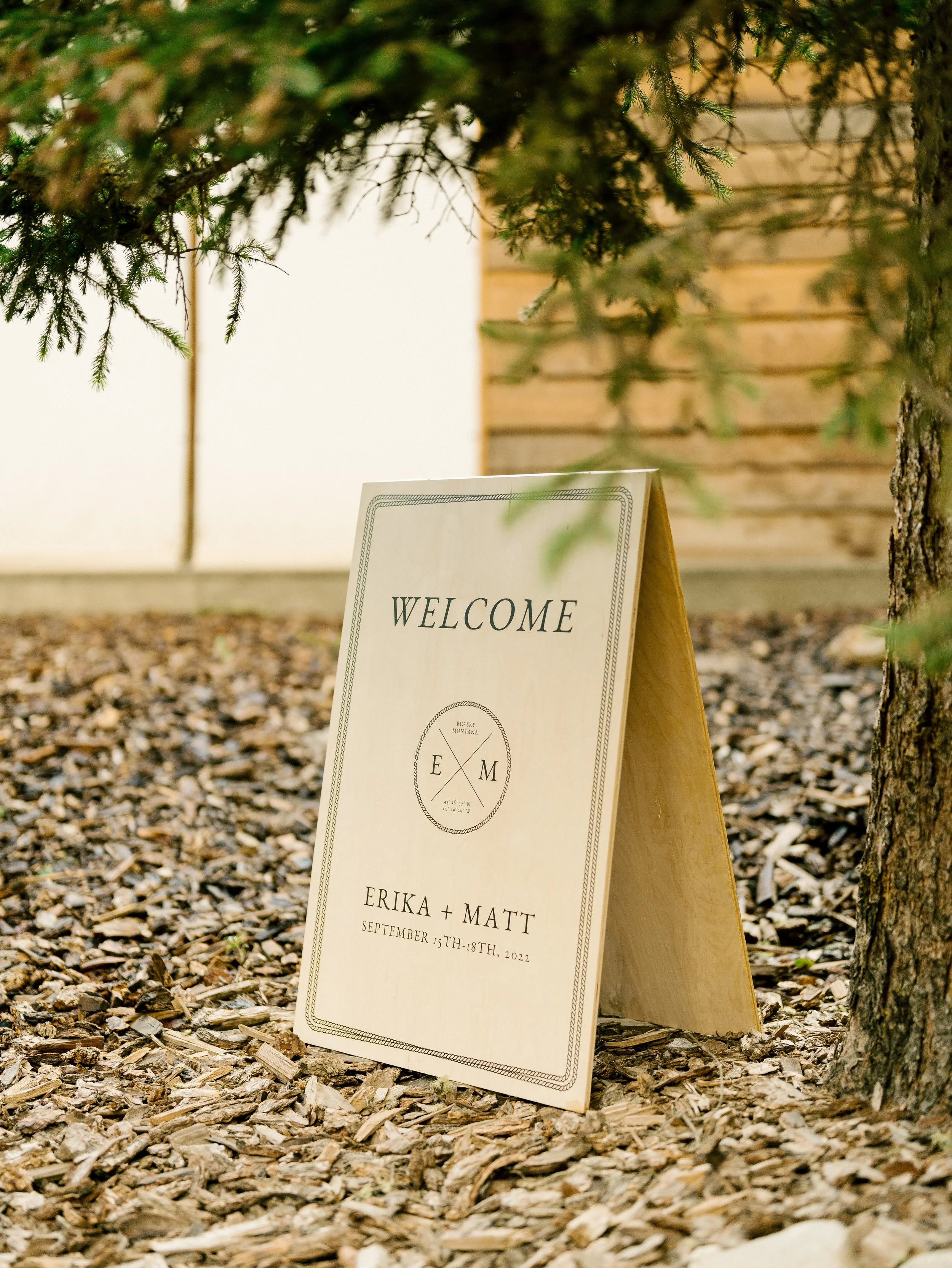 Wedding welcome sign with the names Erika and Matt, dated September 15-18, 2022, placed on a stand outdoors with a background of green ferns and a wooden wall.