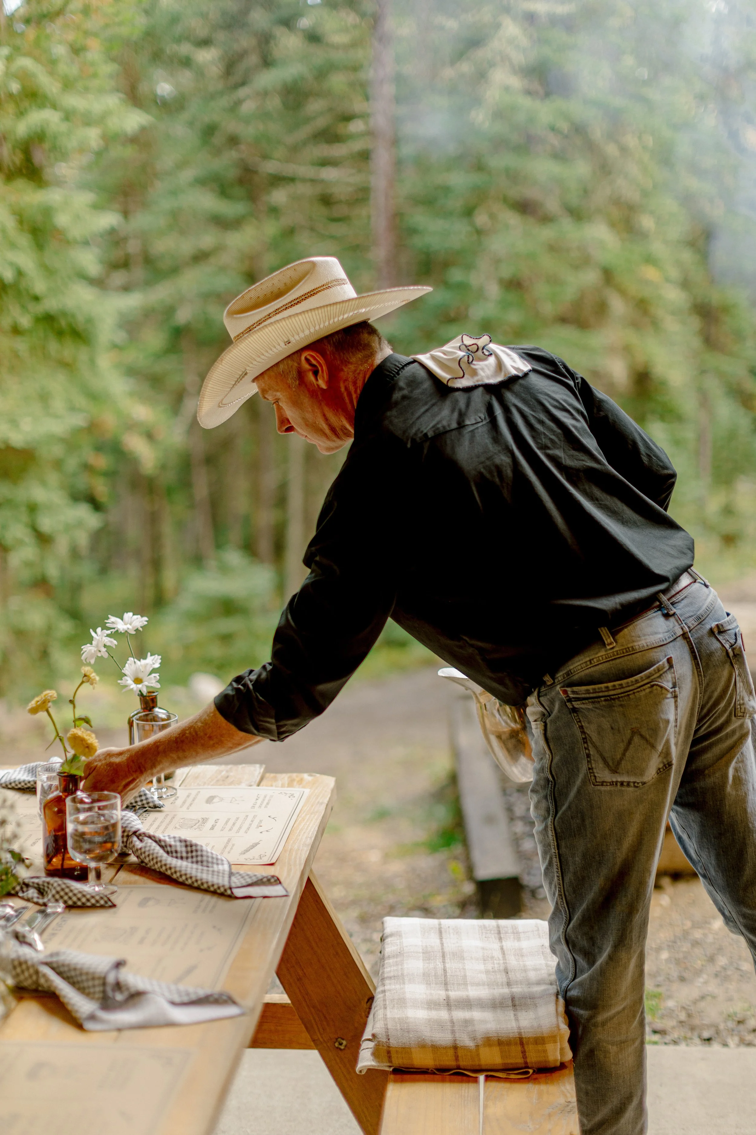 A man wearing a cowboy hat and black shirt setting up a table outdoors with glass vases, flowers, and paper menus.