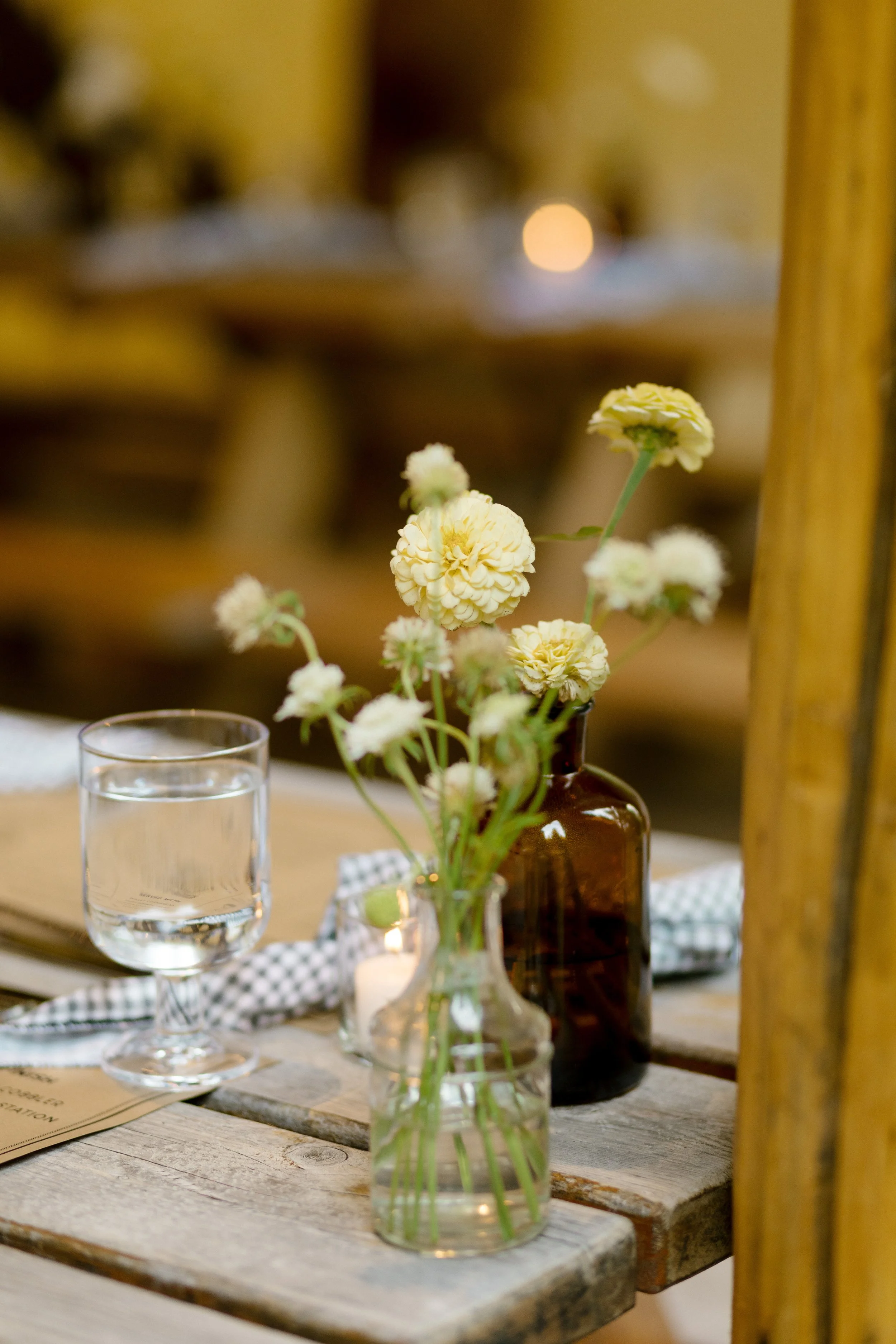 Close-up of a small glass vase holding white and yellow flowers on a rustic wooden table. A glass of water and a menu are nearby, with a warm, softly lit background.