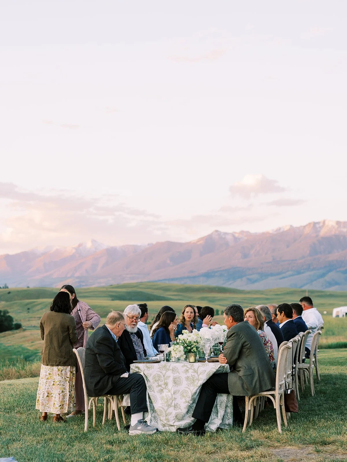 People seated at a long outdoor dining table in a scenic rural landscape with mountains and green fields, during a gathering or celebration.
