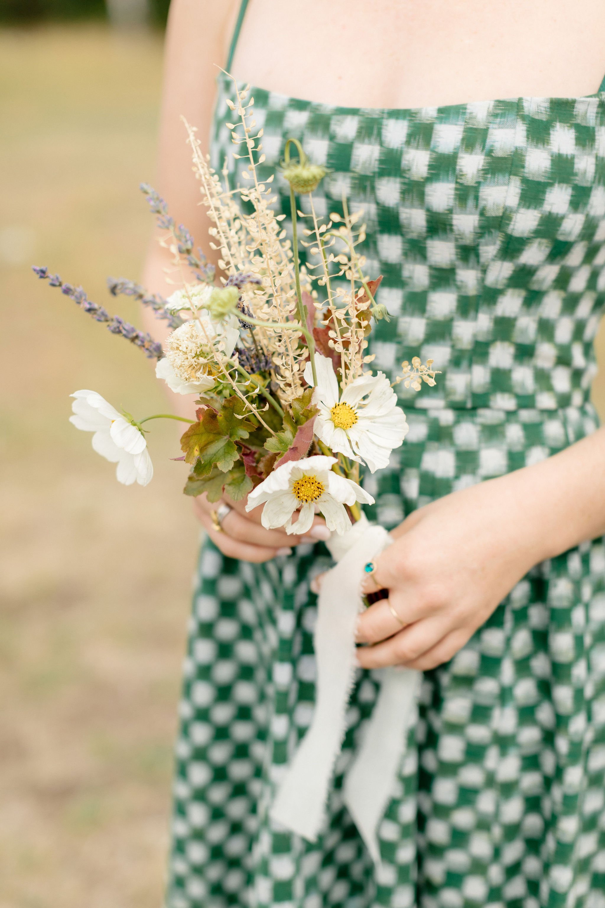 A woman in a green and white checkered dress holding a small bouquet of wildflowers, including white daisies and lavender, with a blurred outdoor background.