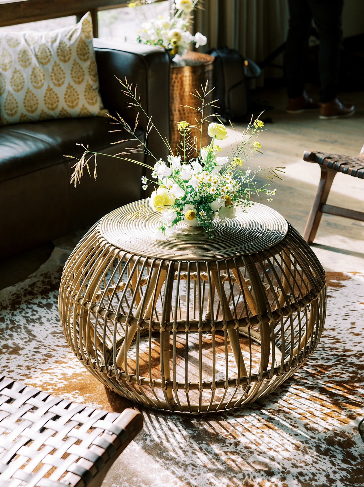 A round, woven rattan coffee table with a decorative top, topped with a floral arrangement in a white vase containing white and yellow flowers, situated in a bright living room with a dark leather sofa, a patterned pillow, a wooden chair, and natural