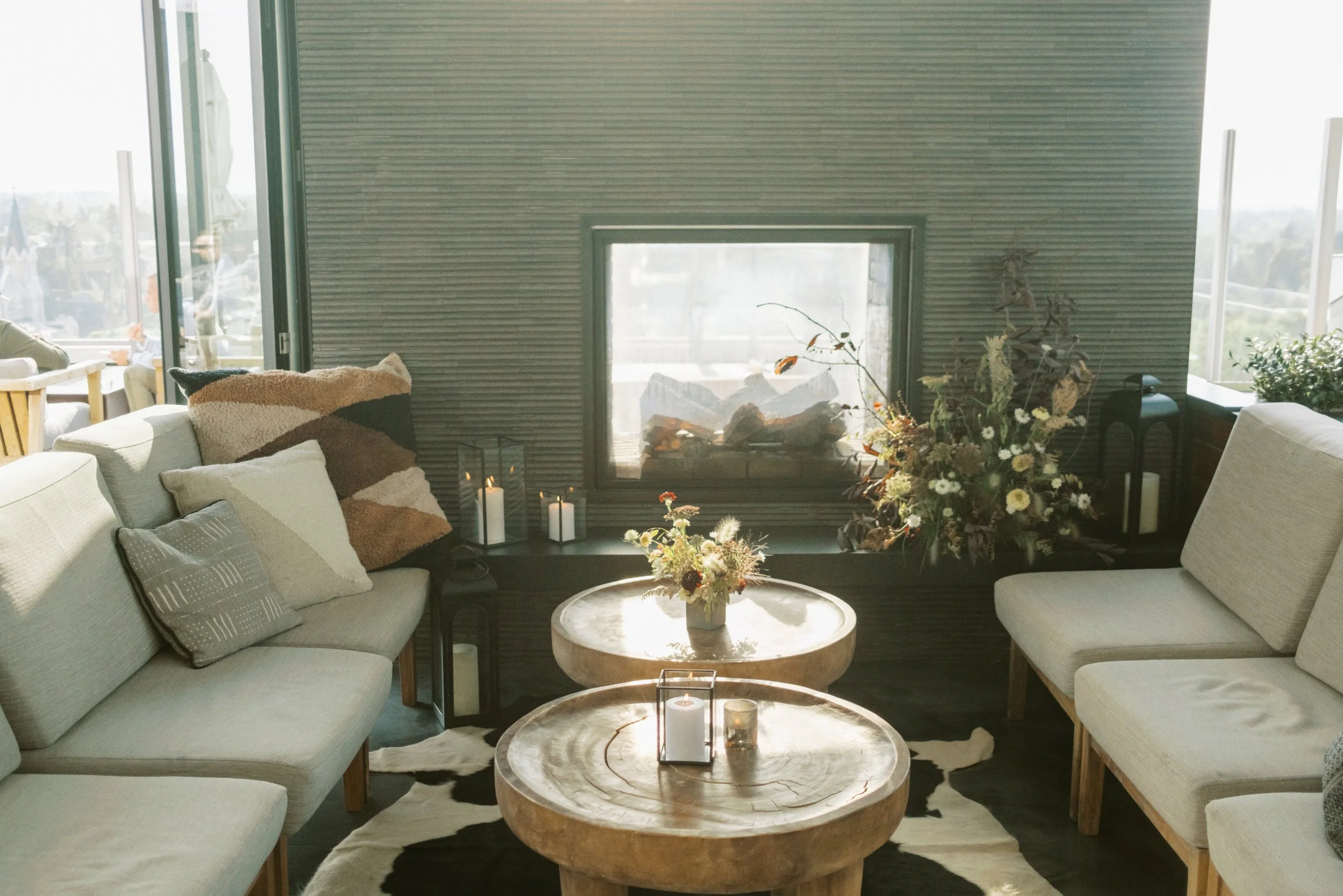 A cozy living room with beige sofas, a round coffee table with candles and a floral arrangement, a fireplace, and large windows allowing natural light in.