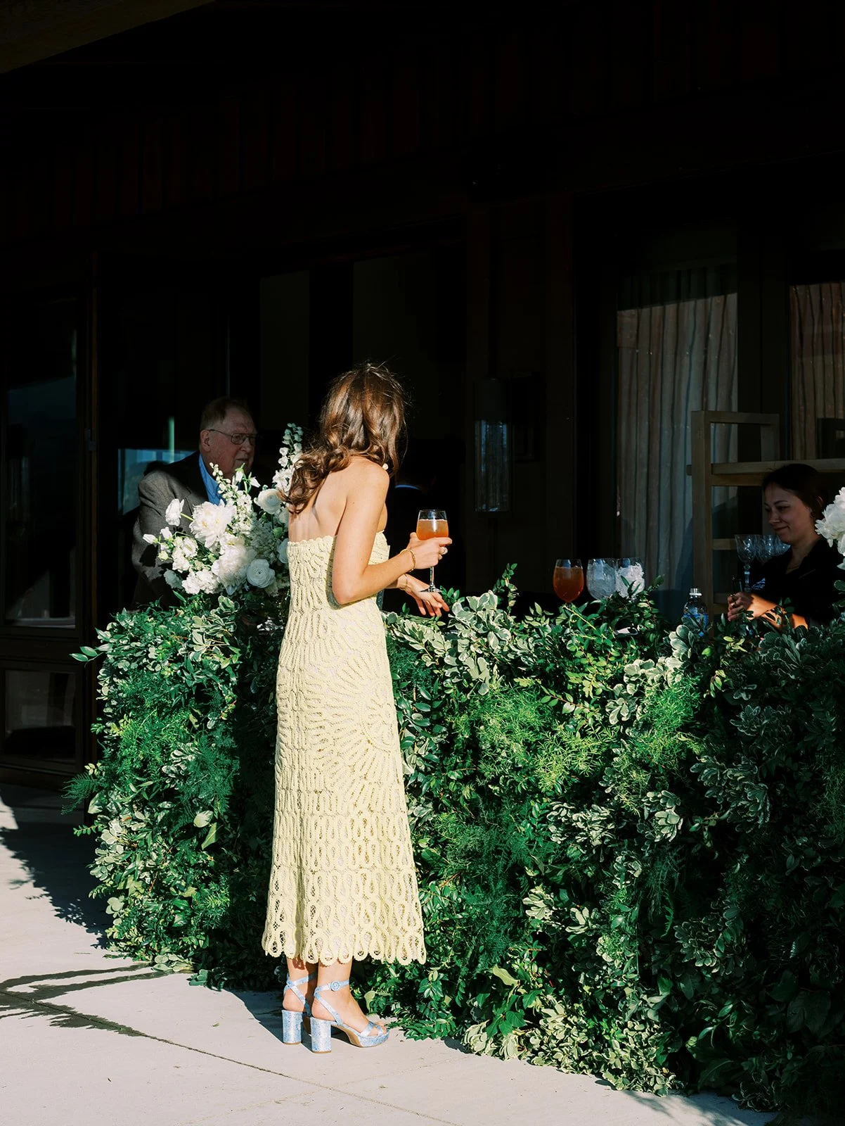 Woman in a yellow dress holding a drink, standing near a bar decorated with green foliage, with two people serving drinks behind the bar.