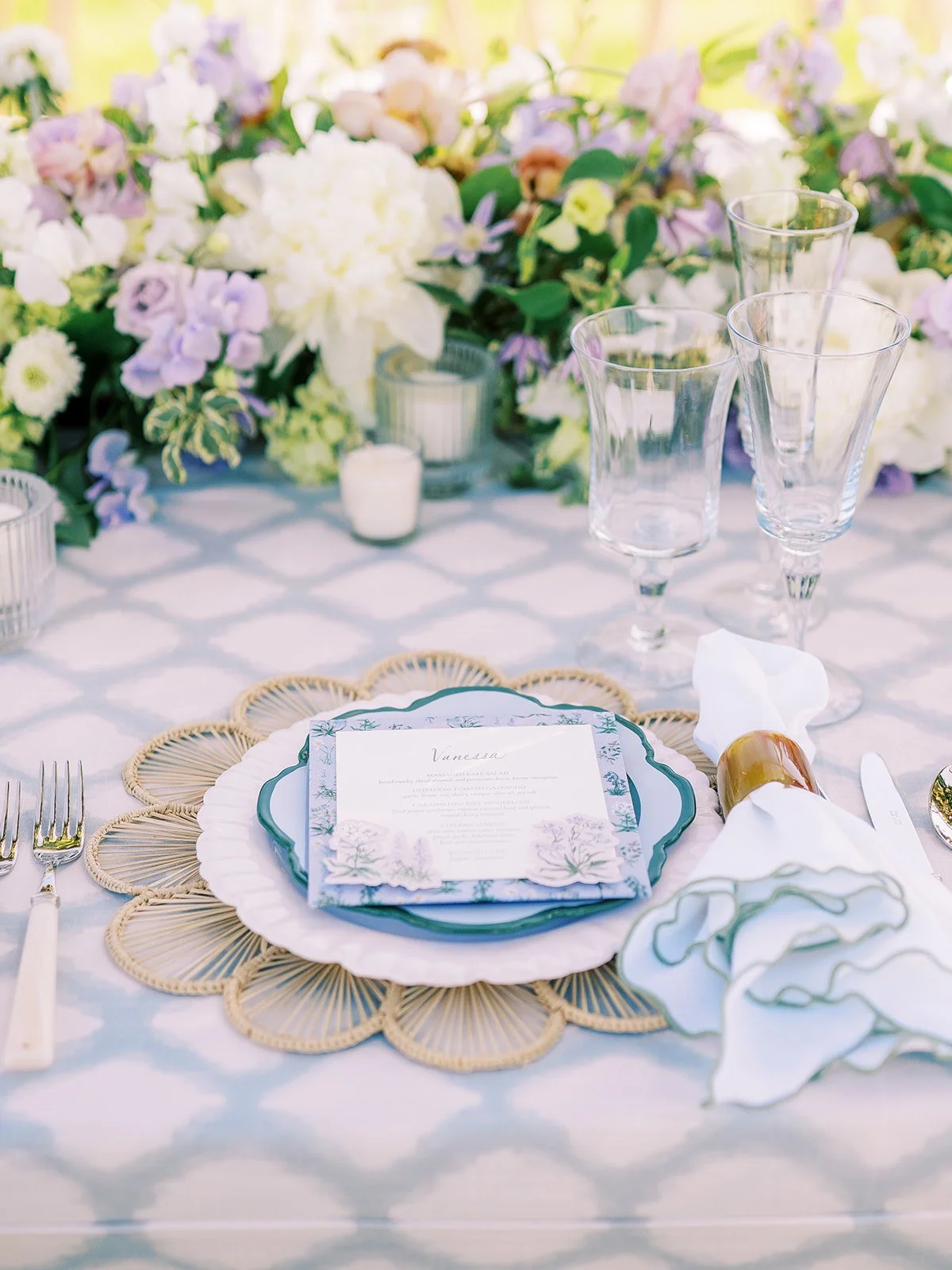 Elegant table setting with floral centerpiece, clear glassware, a menu card, white napkin with gold napkin ring, cutlery, and decorative placemats on a table with a patterned tablecloth.
