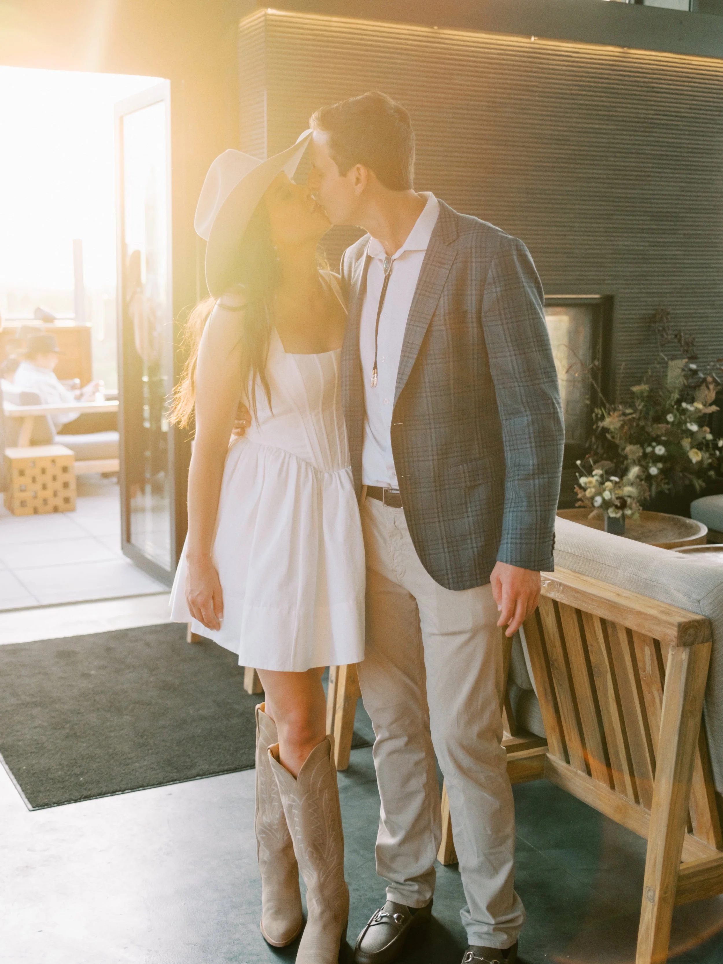 A couple sharing a kiss indoors, with sunlight streaming through a window, in a modern living space with a sofa, flower arrangement, and wooden furniture.