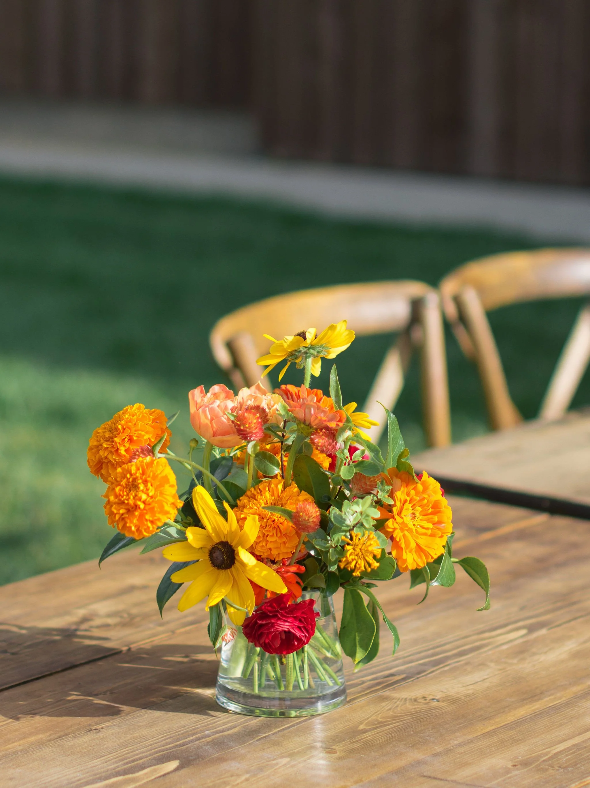 Bright sunflower and marigold floral arrangement in warm orange and yellow tones on a wooden surface, by Wild Blume