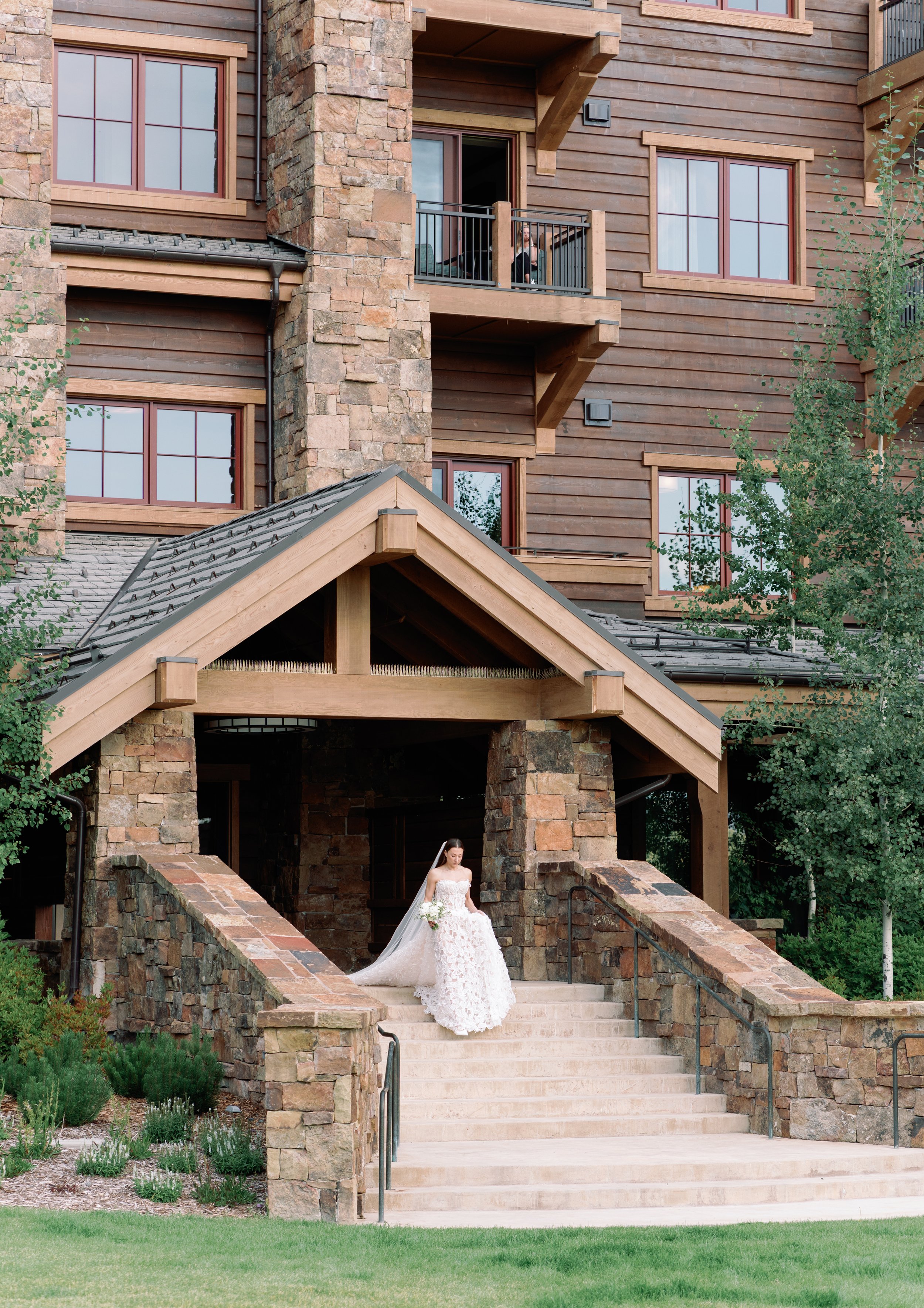 A bride in a white wedding dress with a long train walking down stone steps in front of a multi-story wooden and stone house with balconies and large windows.