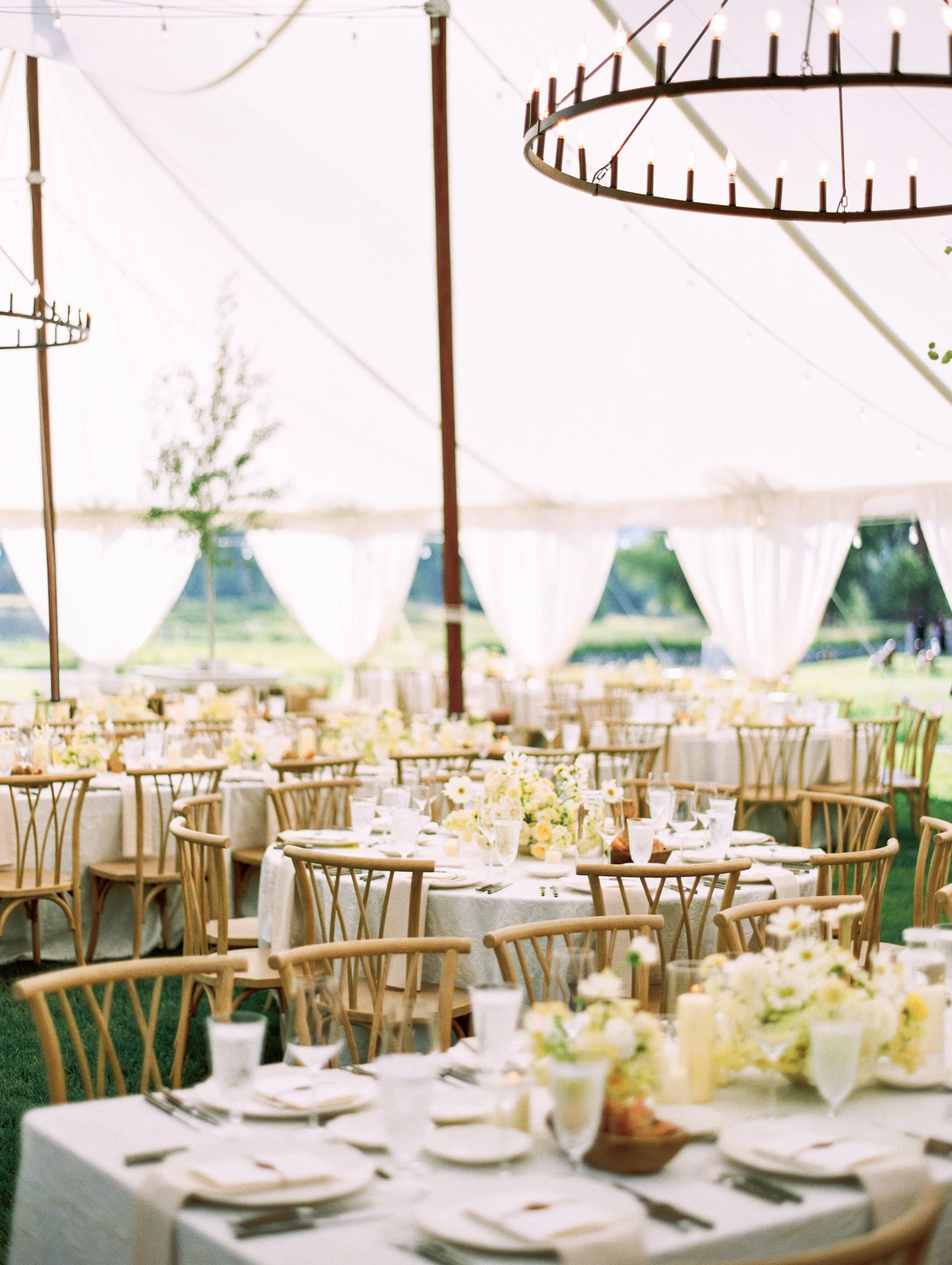Elegant tented wedding reception with round dining tables, white floral centerpieces, draped fabric, and warm overhead lighting, designed by Wild Blume