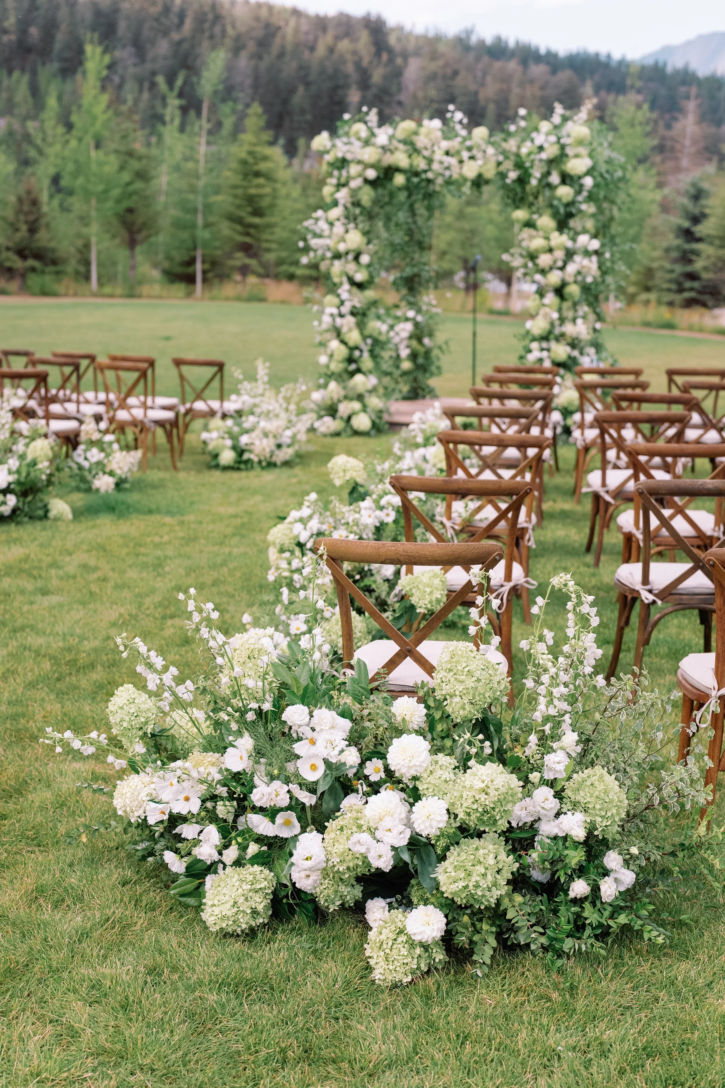 Outdoor wedding setup with a floral arch and chairs on a grassy field, surrounded by trees and mountains in the background.