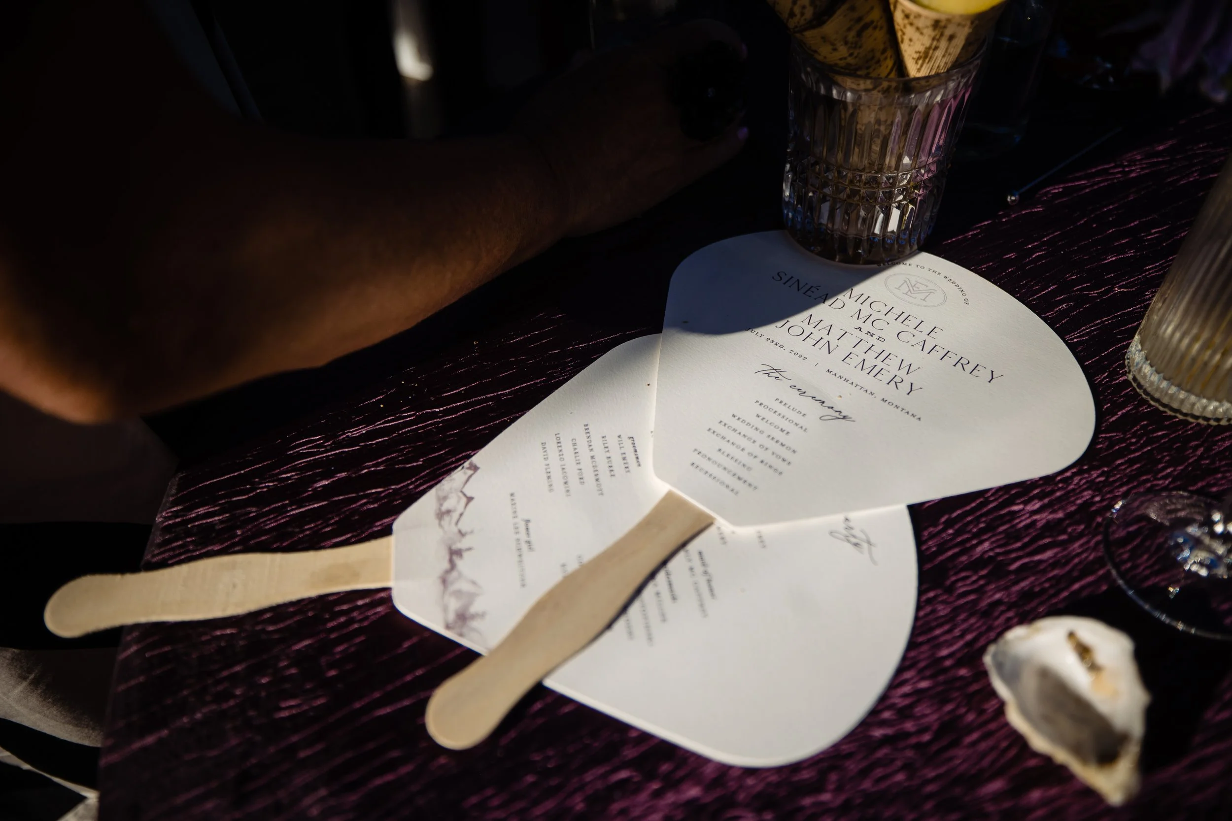 A wedding invitation on a dark purple textured surface with a glass containing yellow rolled-up papers, a hand with rings, and various small objects around.