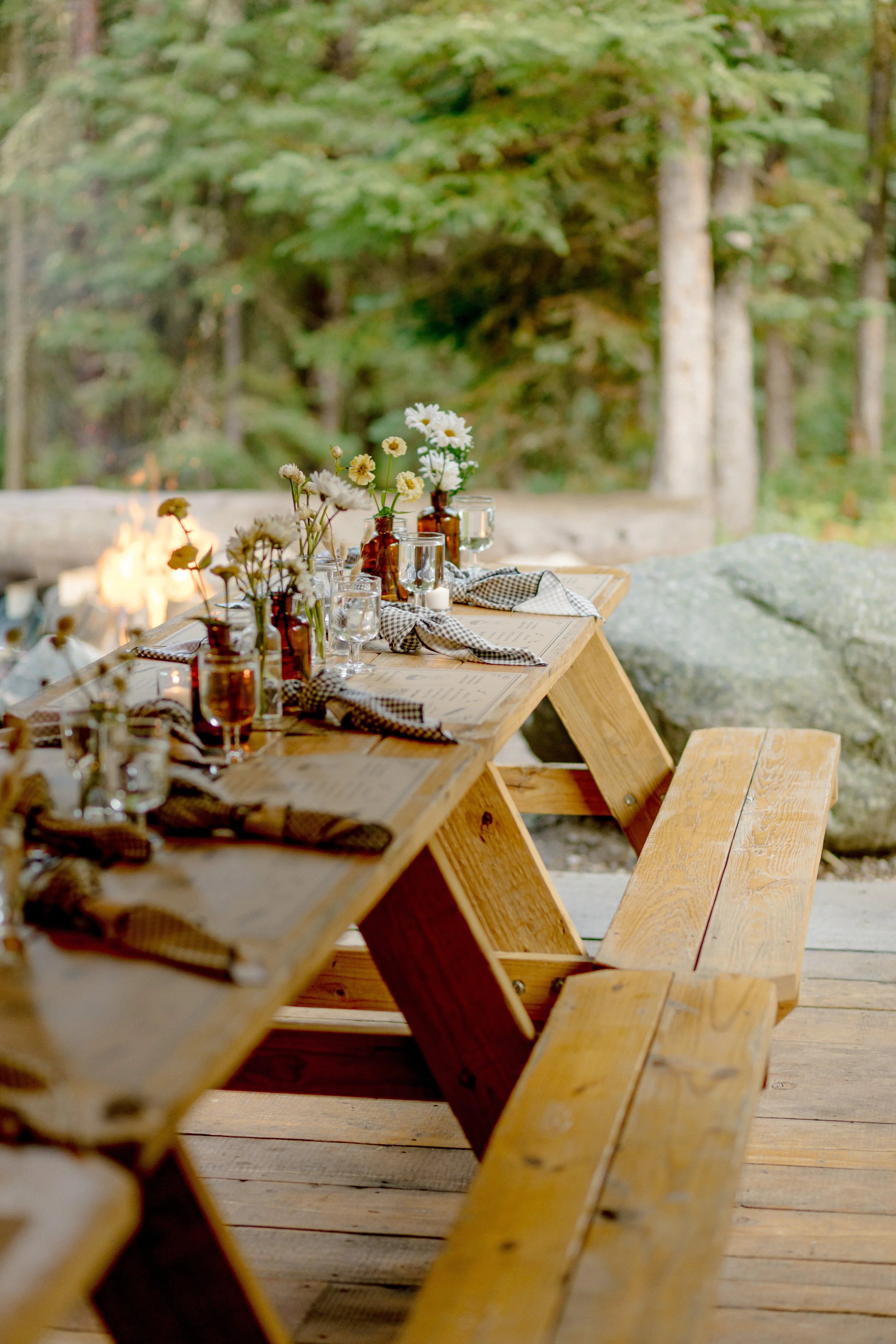A wooden outdoor dining table set with vases of white and yellow flowers, glassware, and checkered napkins, situated in a forest environment.