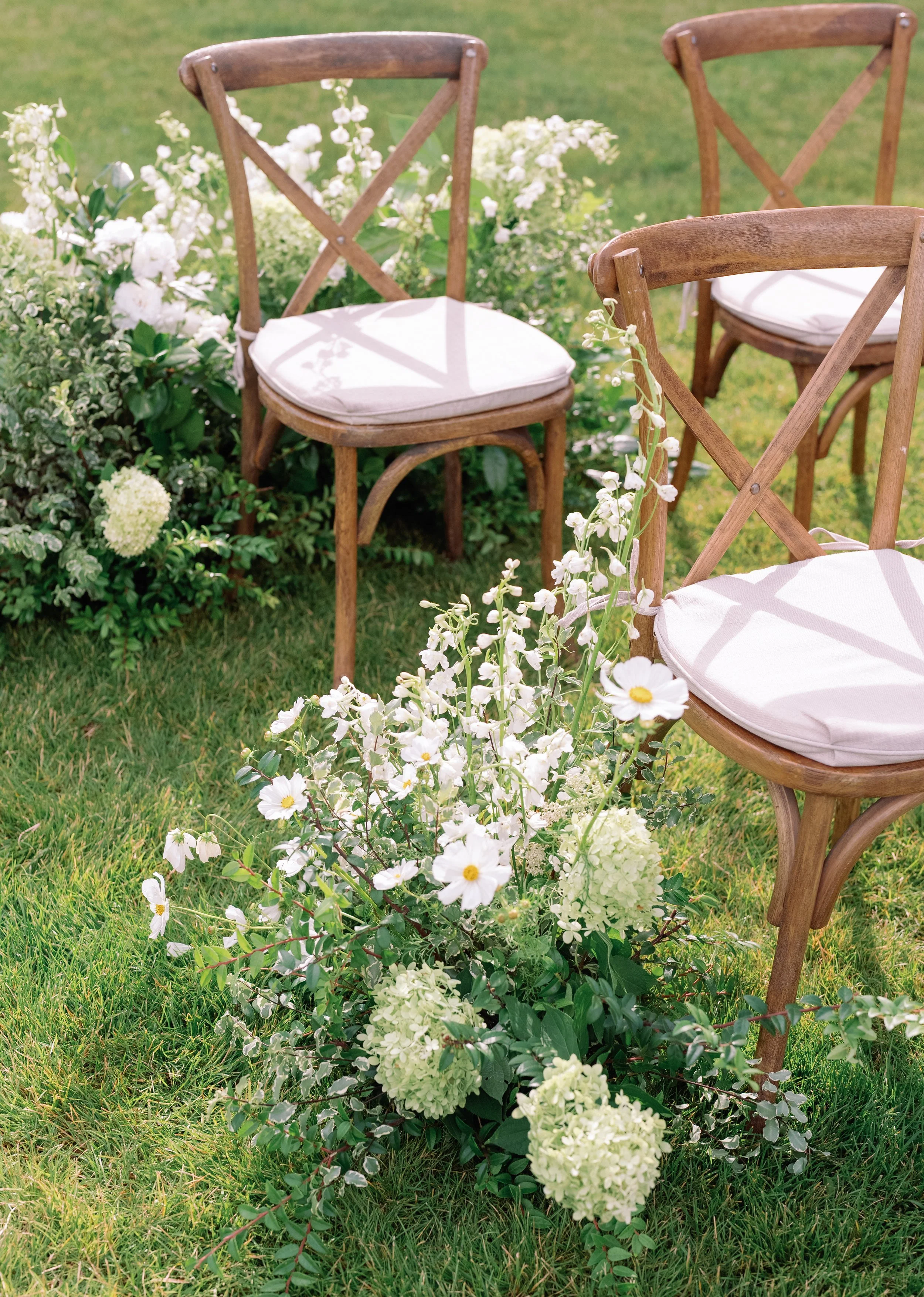 Outdoor scene with wooden chairs and white cushions surrounded by white and green flowers, on a grassy lawn.