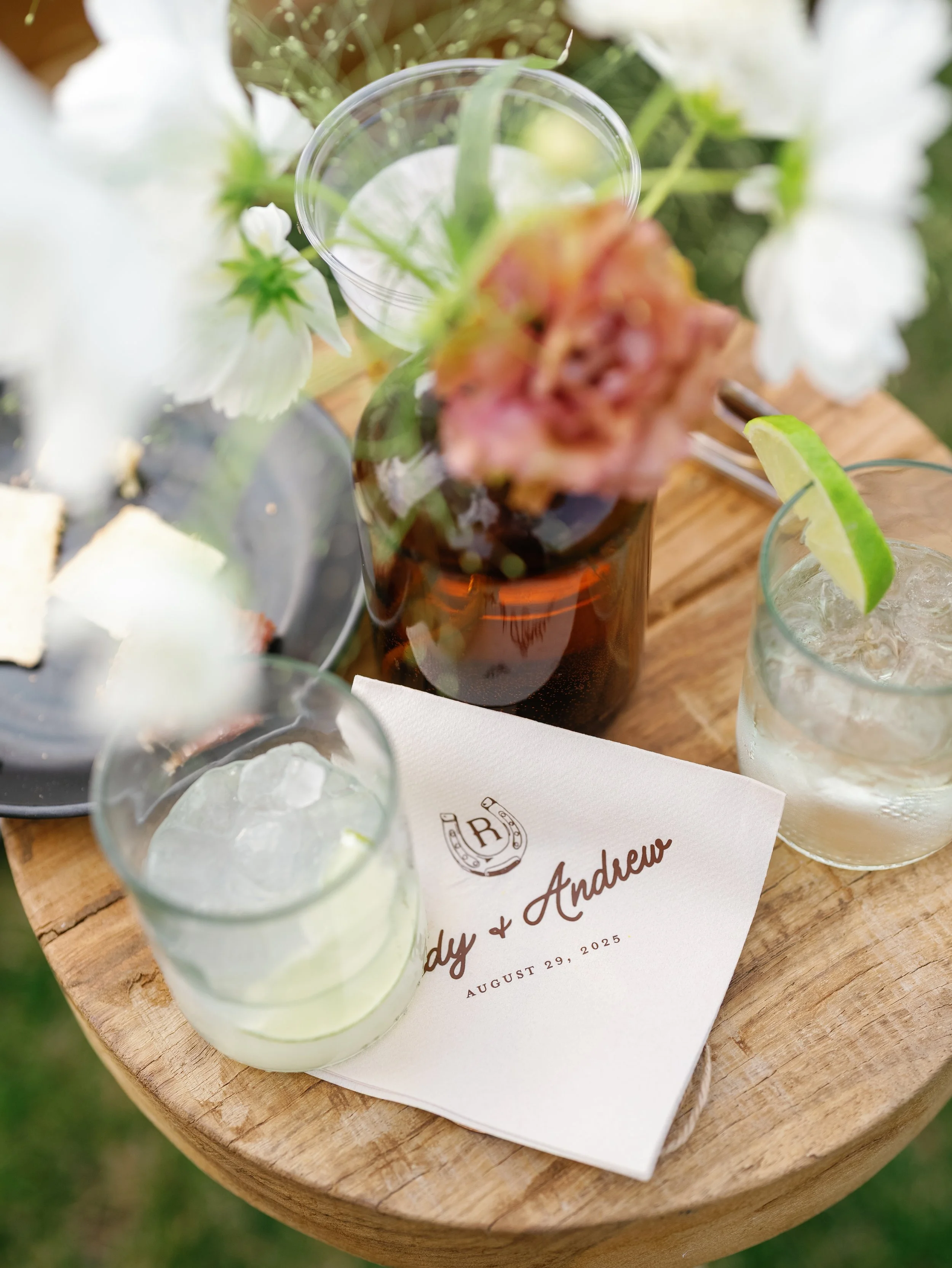 Close-up of drinks on a wooden table, with a white card displaying the names 'Andy & Andrew' and the date August 29, 2025, at a wedding celebration.