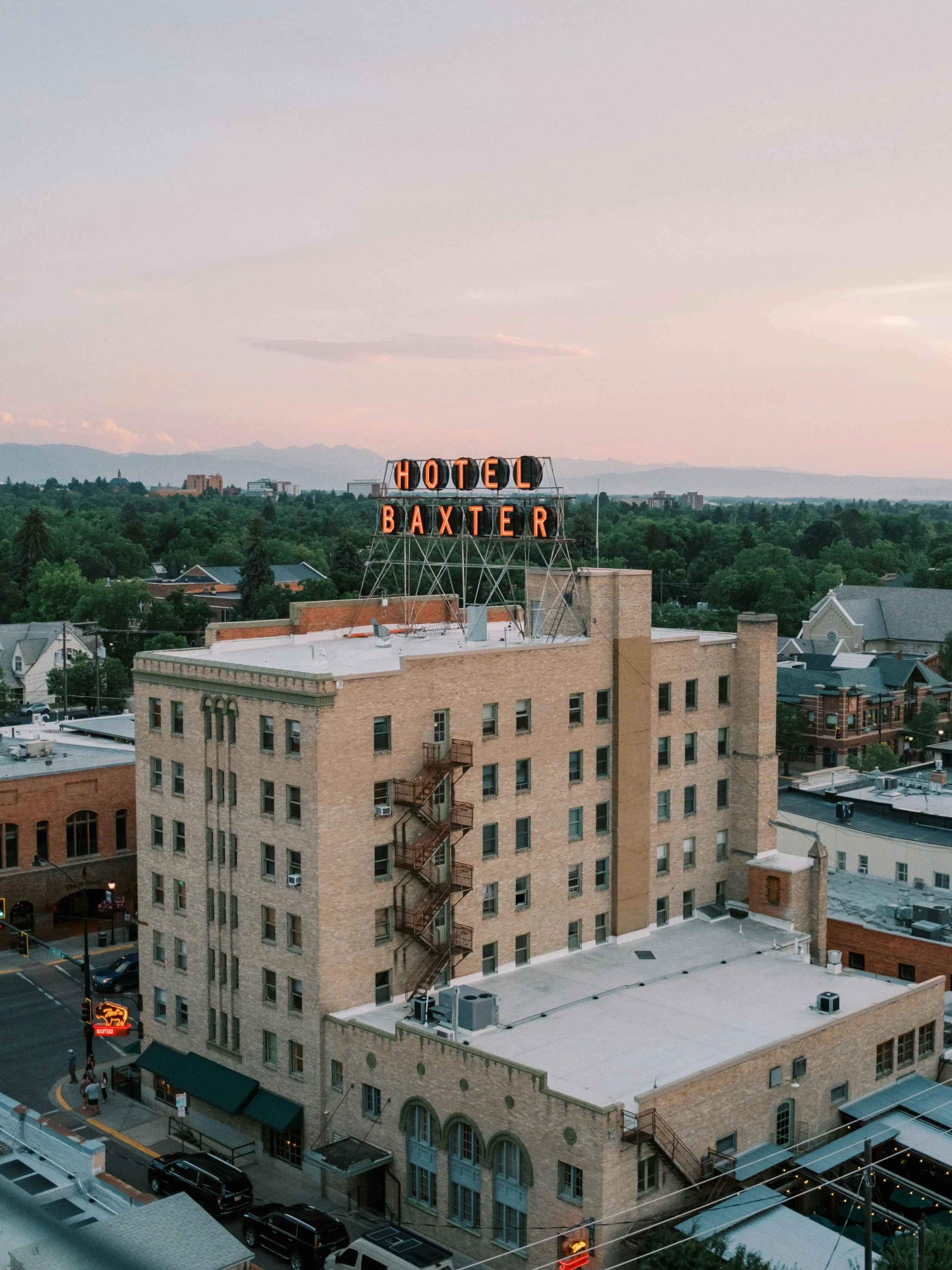 A mid-rise brick building with a neon sign on the roof that reads 'Hotel Baxter'. The building is surrounded by trees and has a fire escape on one side. There are cars parked along the street and a small crowd of people near the sidewalk.