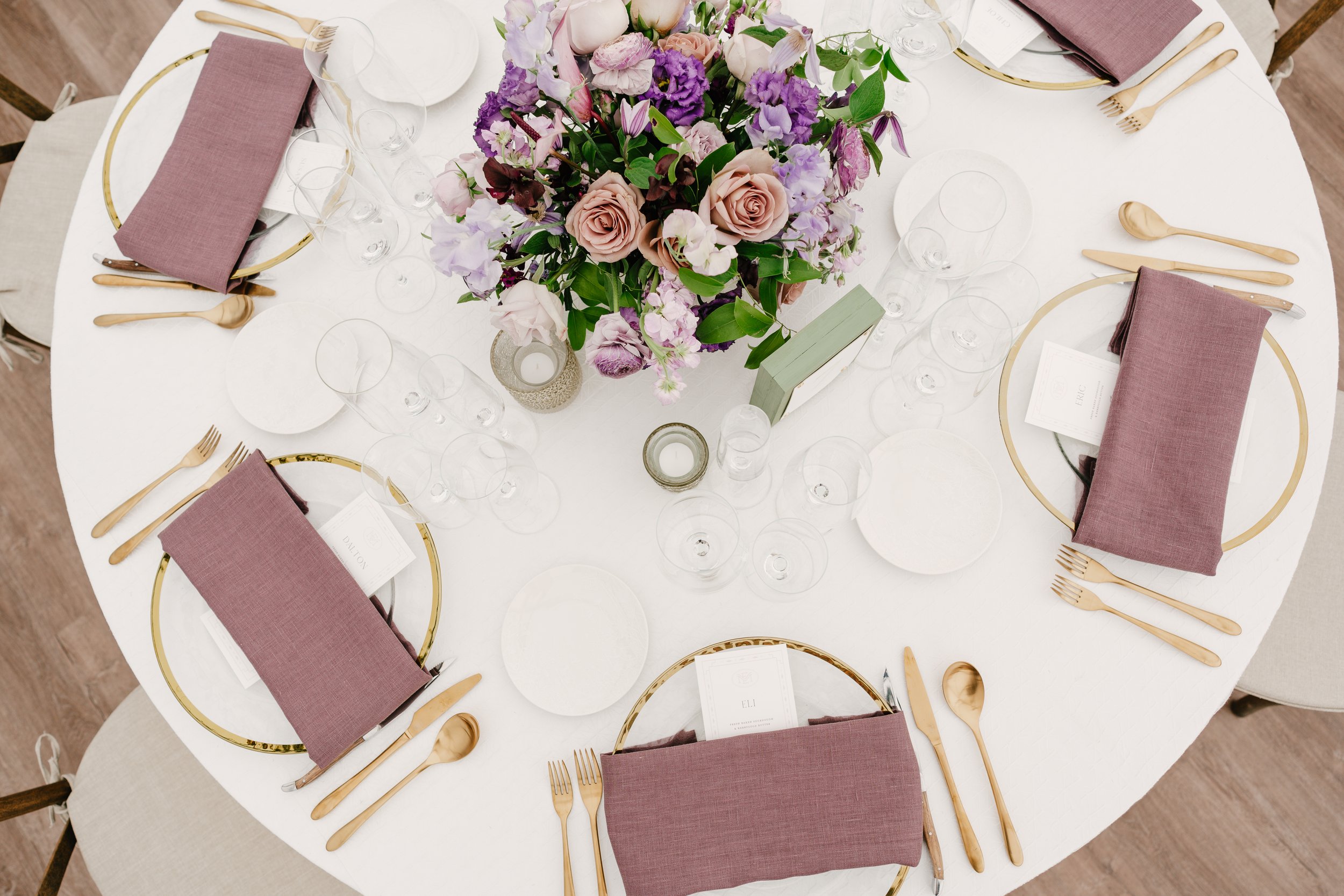 A round table set for a formal dinner with a floral centerpiece of pink and purple flowers, gold-rimmed plates, mauve cloth napkins, gold flatware, and empty glassware arranged around it.