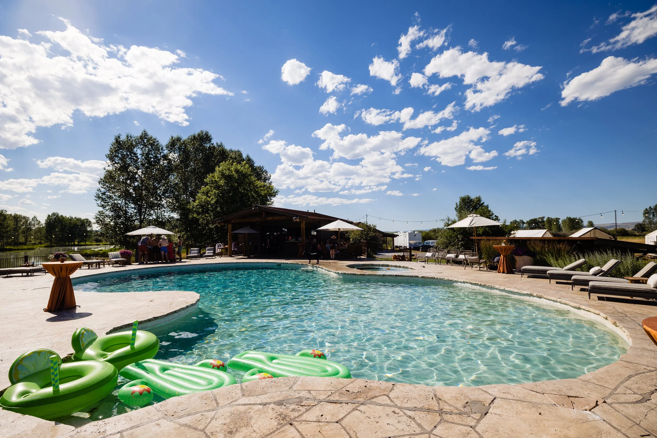 An outdoor swimming pool with green inflatable floaties, lounge chairs, umbrellas, and a building in the background under a cloudy sky.