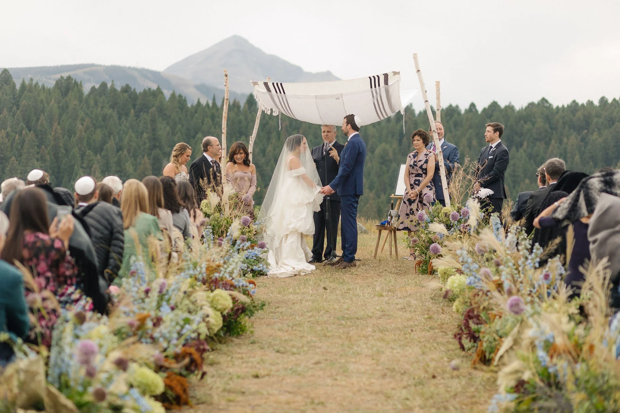 A wedding ceremony outdoors with a bride and groom standing under a makeshift canopy, surrounded by an audience, with mountains and trees in the background, and decorated with colorful flowers along the aisle.