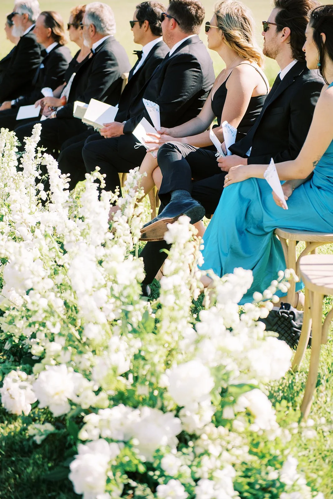 People sitting in chairs during an outdoor wedding ceremony, holding programs, with white flowers in the foreground.
