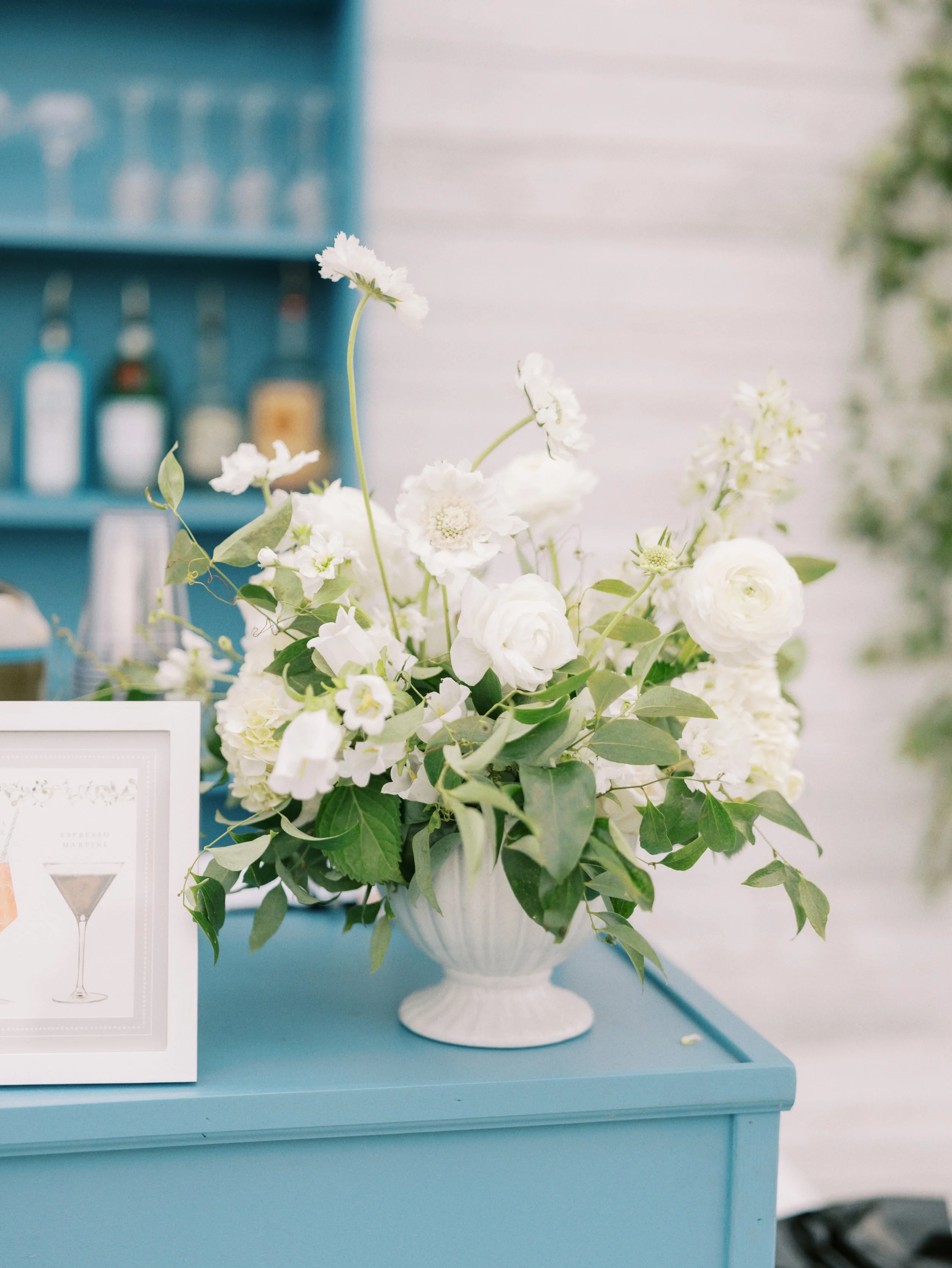 A bouquet of white flowers with green leaves in a white vase on a blue table.