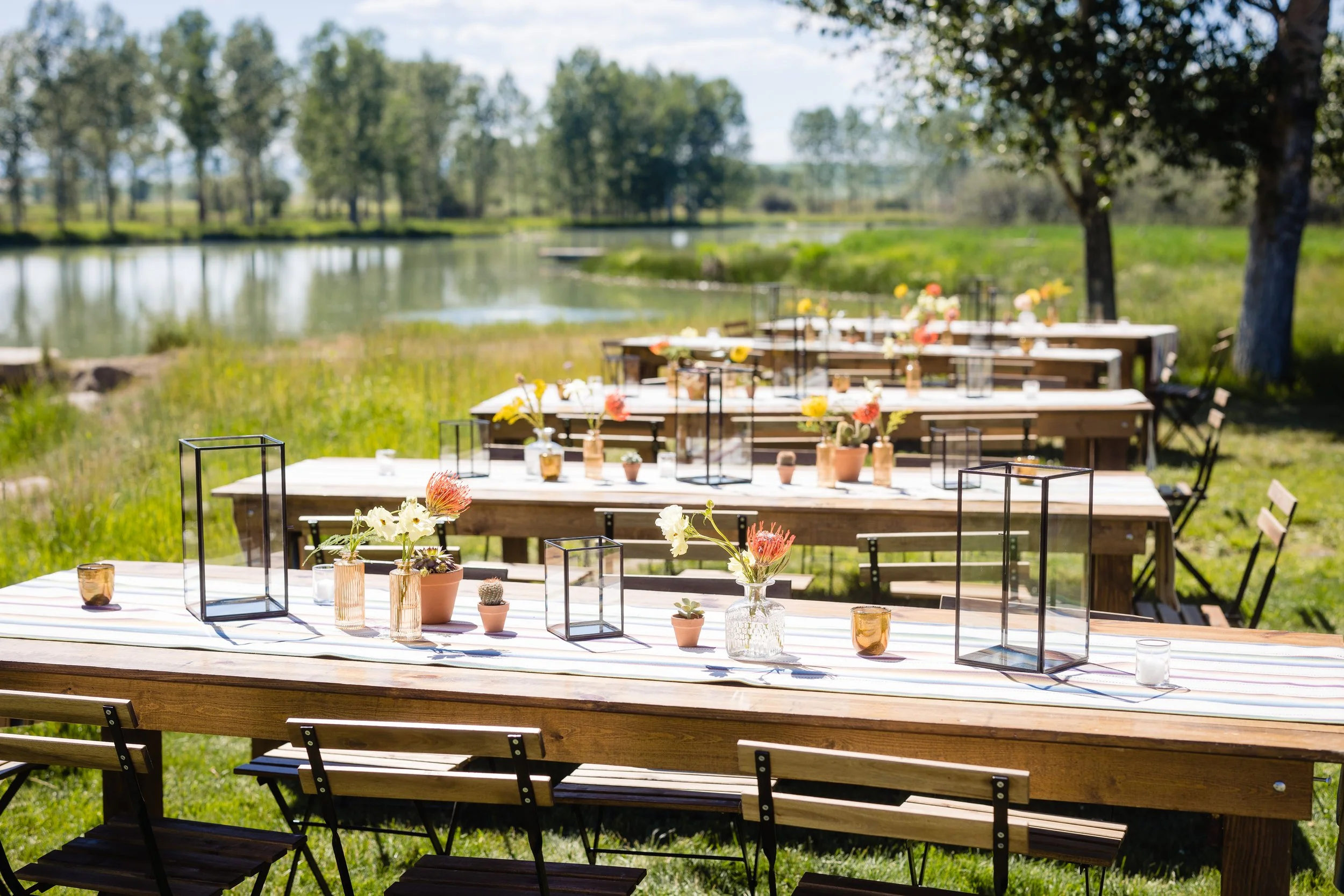 Outdoor celebration setup with long tables decorated with flowers in vases and candle holders near a lake and trees.