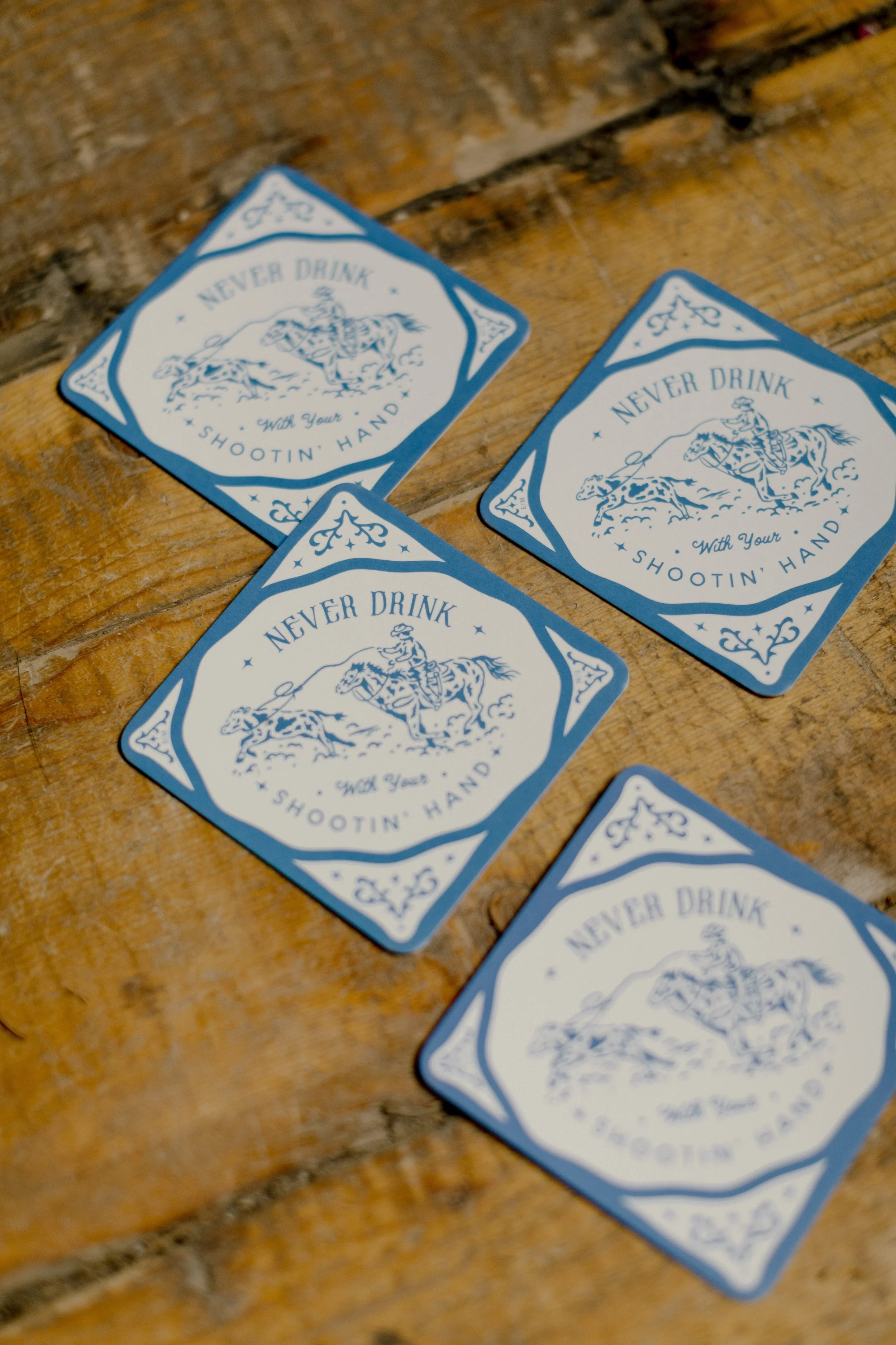 Blue cards with a cowboy riding a bucking horse, with the text "Never Drink With Your Shootin' Hand" on a wooden surface.