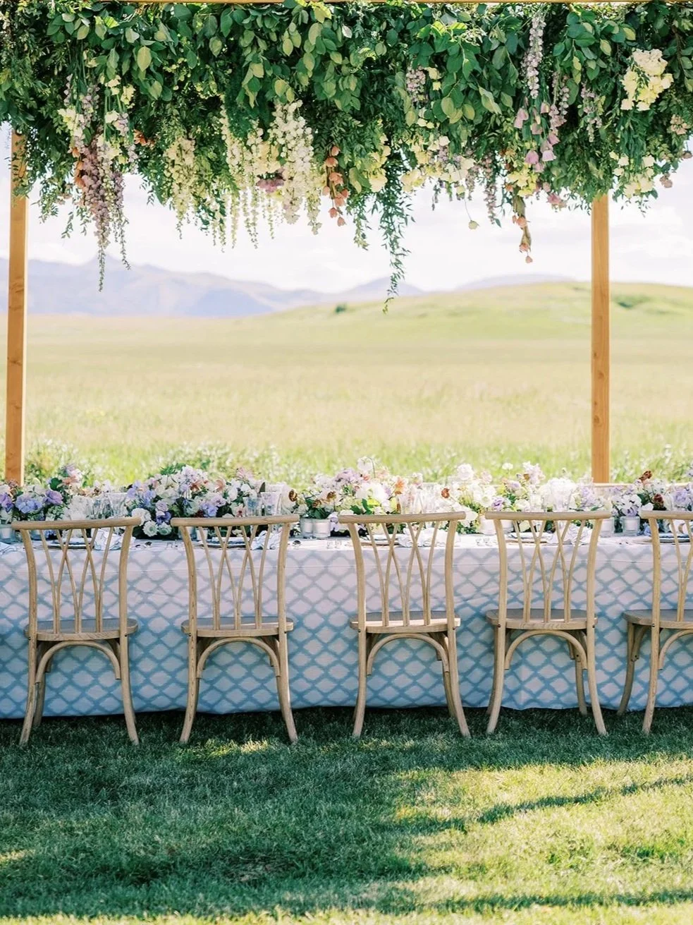 Outdoor lakeside wedding ceremony with lush greenery and white floral arch overhead, rows of wooden chairs, designed by Wild Blume