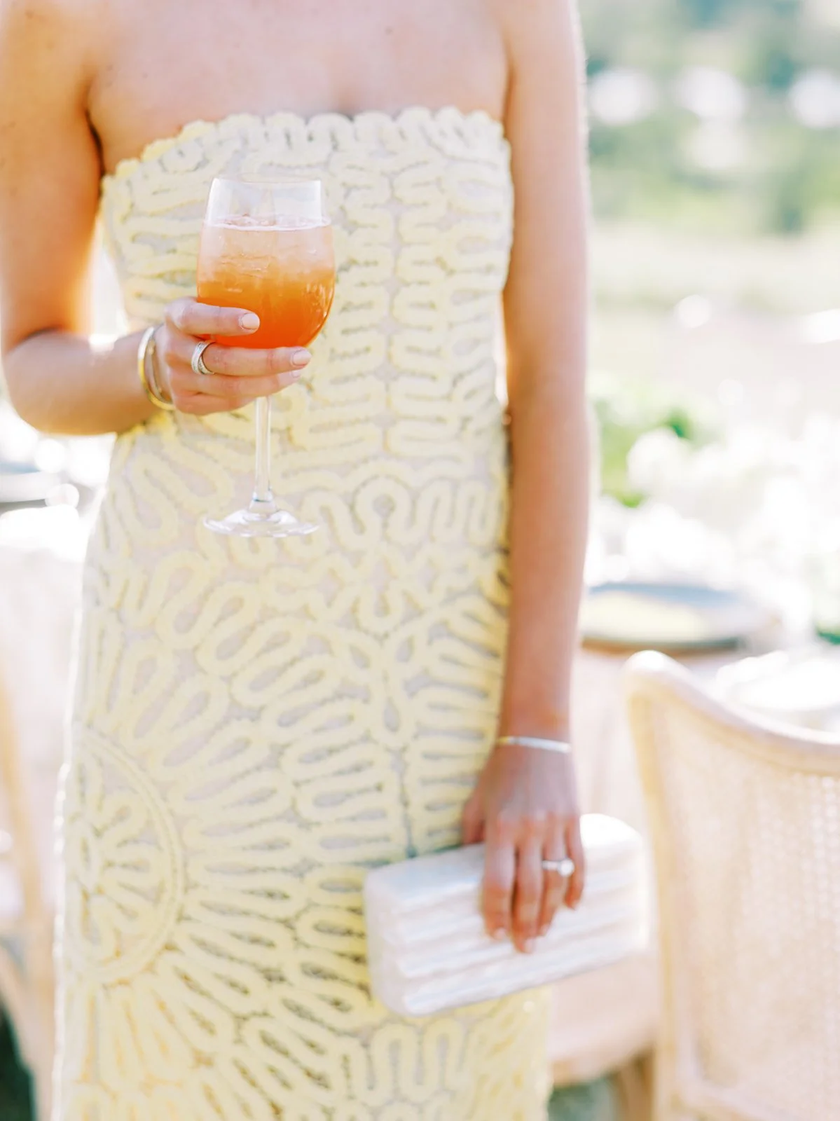 A woman in a yellow and white patterned strapless dress is holding a glass of pink cocktail in her right hand and a white clutch purse in her left hand.