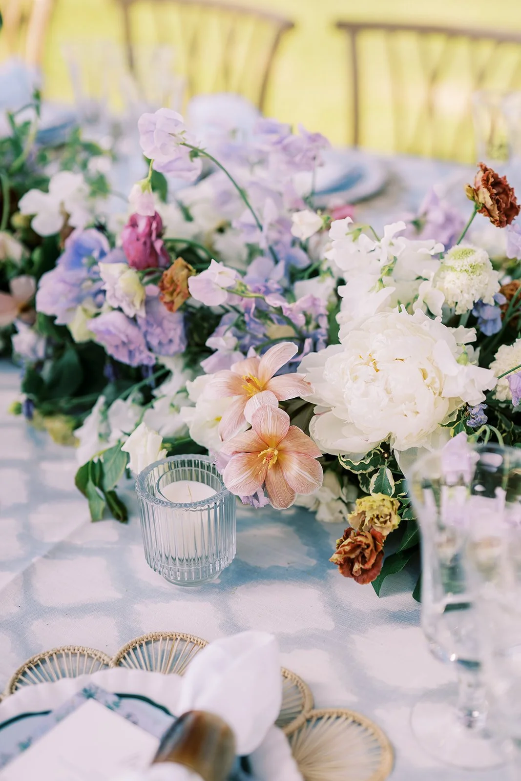 A floral centerpiece with purple, white, pink, and brown flowers on a table, illuminated by a candle in a glass holder, with blurred chairs in the background.