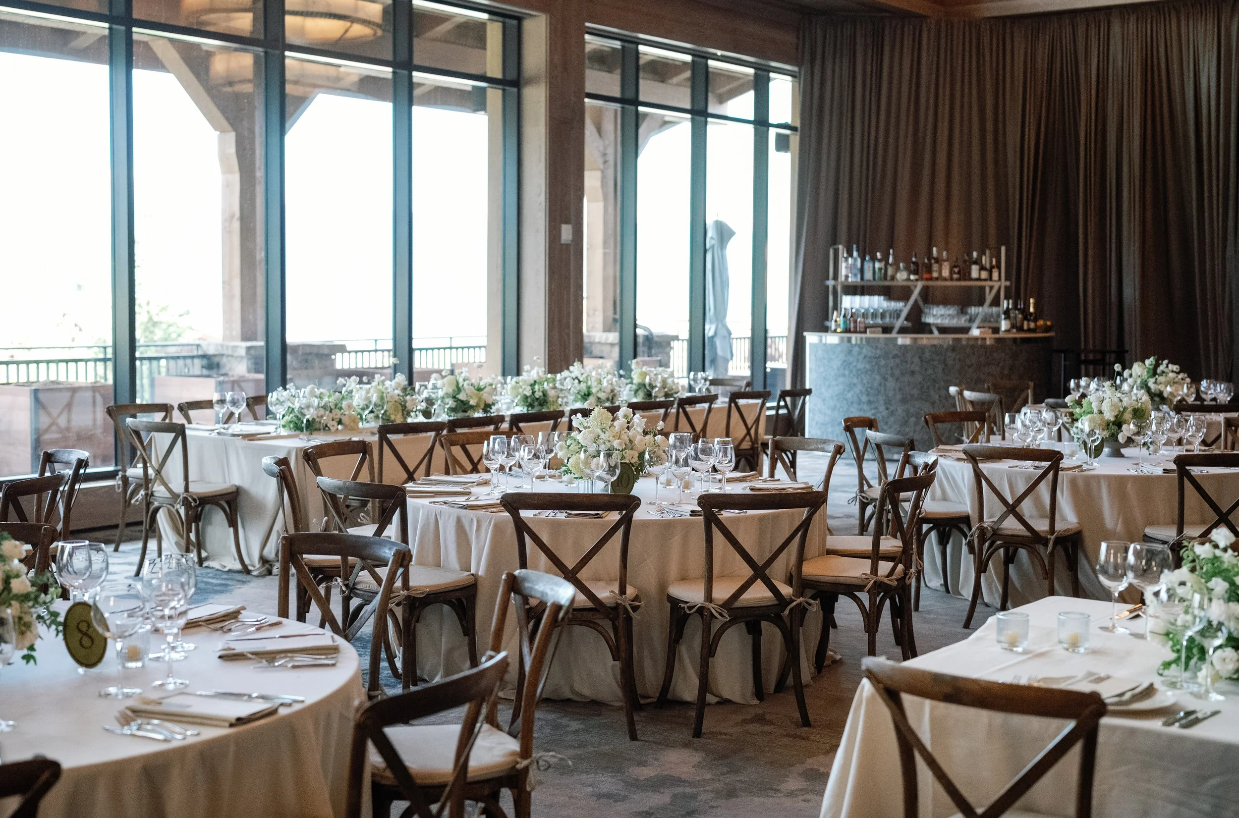 Interior of a decorated banquet hall with round tables covered in white tablecloths, set with glassware, silverware, and floral centerpieces, large windows, and a bar area with bottles and glasses.